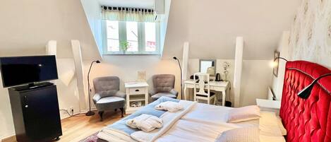 Une chambre d’hôtel avec un grand lit, deux fauteuils, un bureau avec un ordinateur, une télévision et un petit meuble de rangement.
