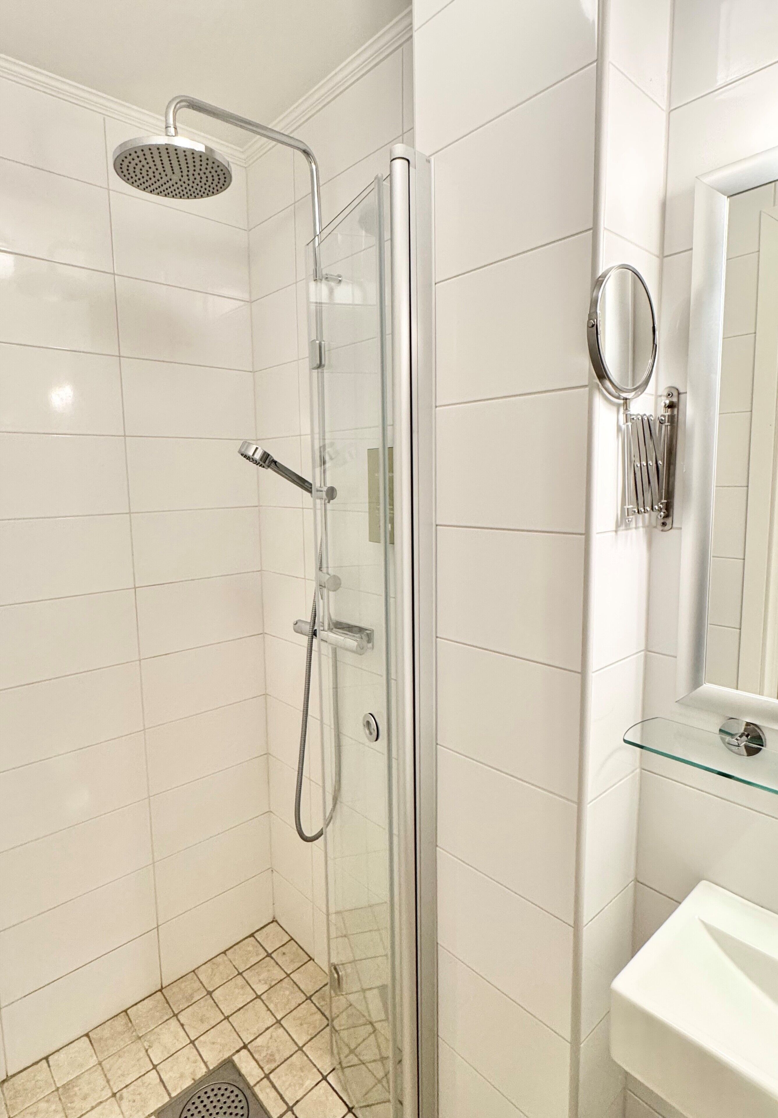 Um chuveiro com box de vidro, um chuveiro manual, uma pia de vidro com espelho e piso revestido de azulejos.