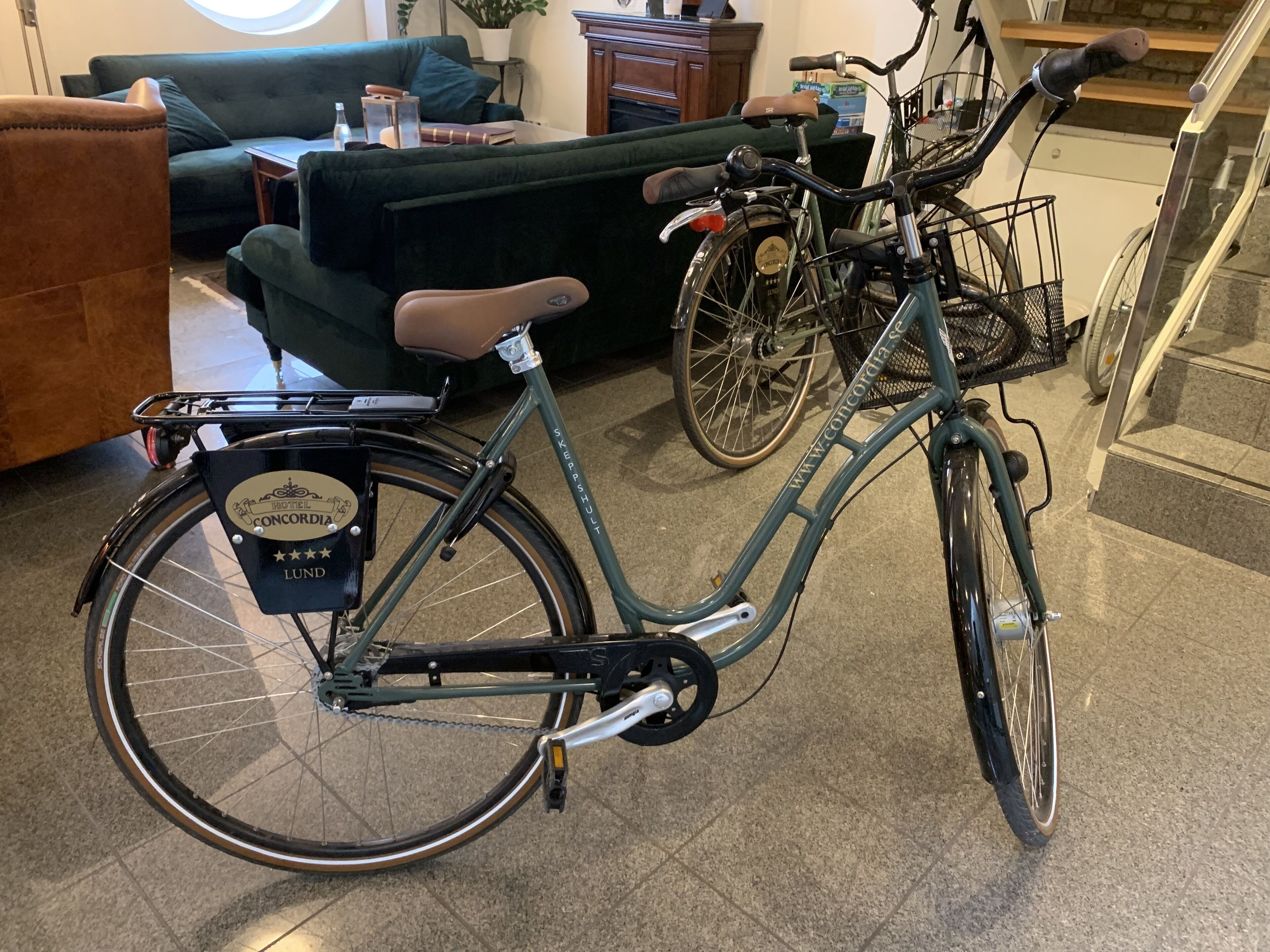 Una bicicleta color turquesa con una canasta delantera y un portaequipajes, estacionada en un interior cerca de una escalera.
