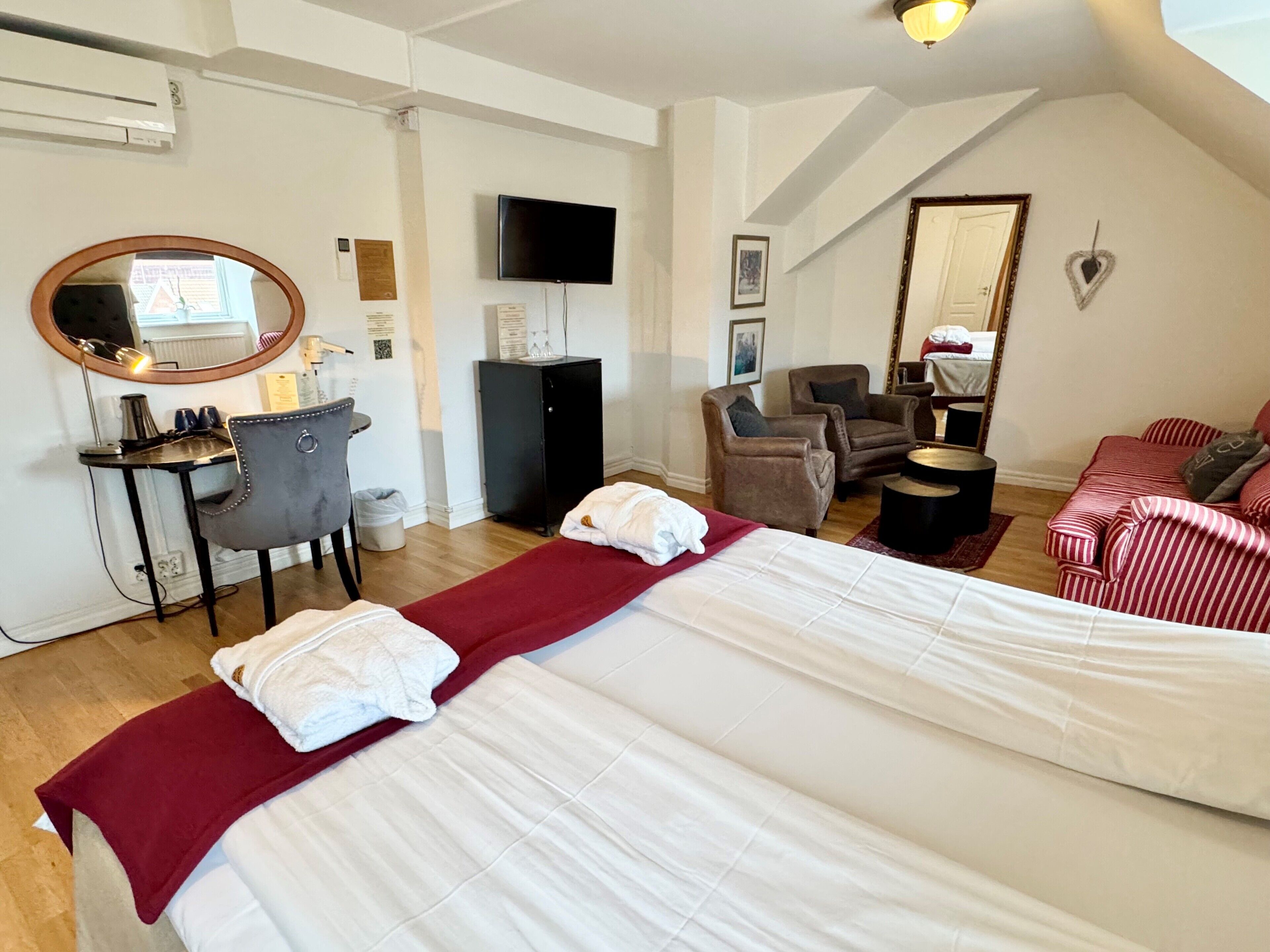 A hotel room with a large bed, a desk with a chair, a television, and a seating area with armchairs and a sofa.