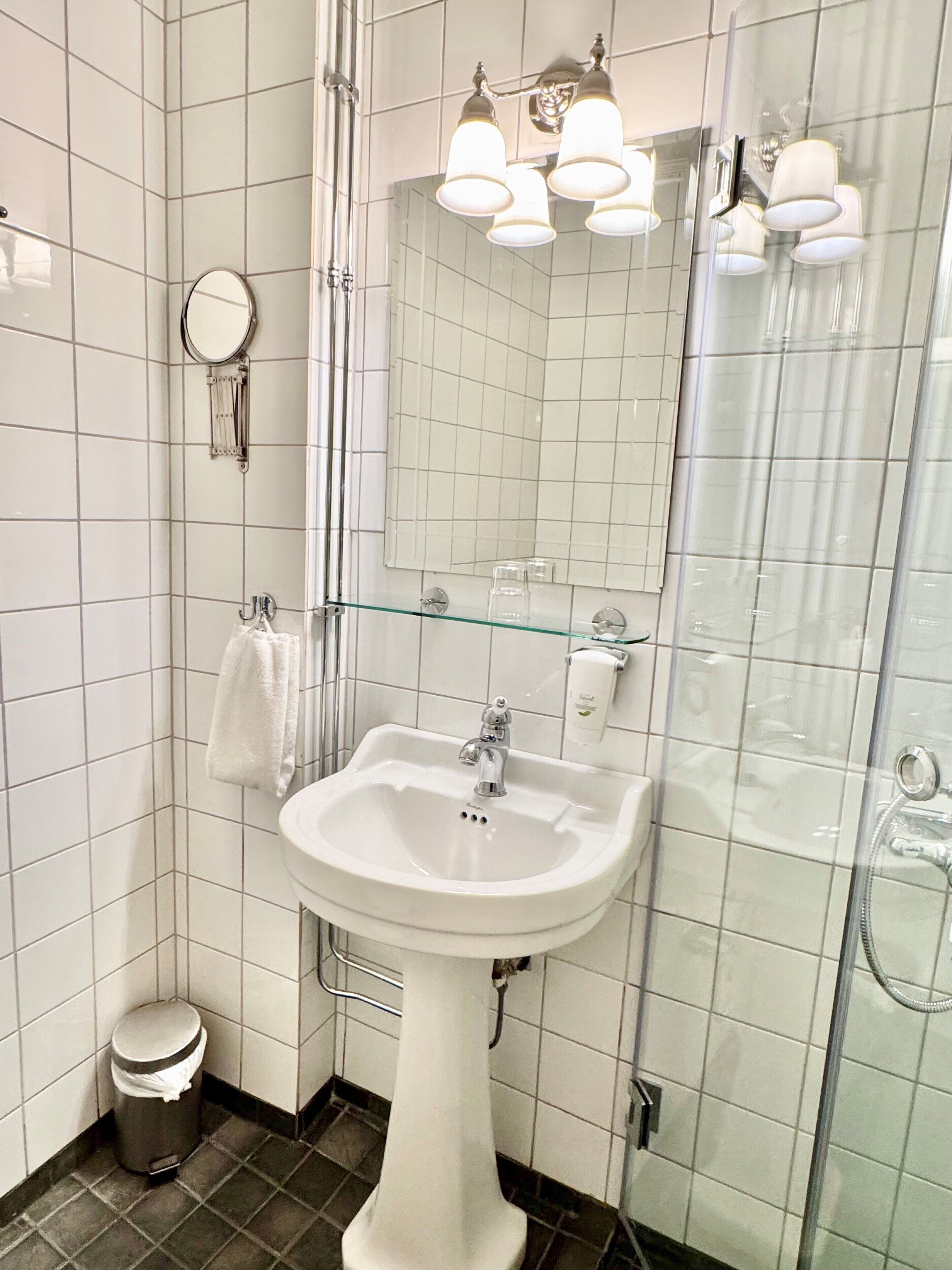 Un baño con paredes de azulejos blancos, un lavamanos blanco, una cabina de ducha con mampara de vidrio y un espejo con marco redondo.