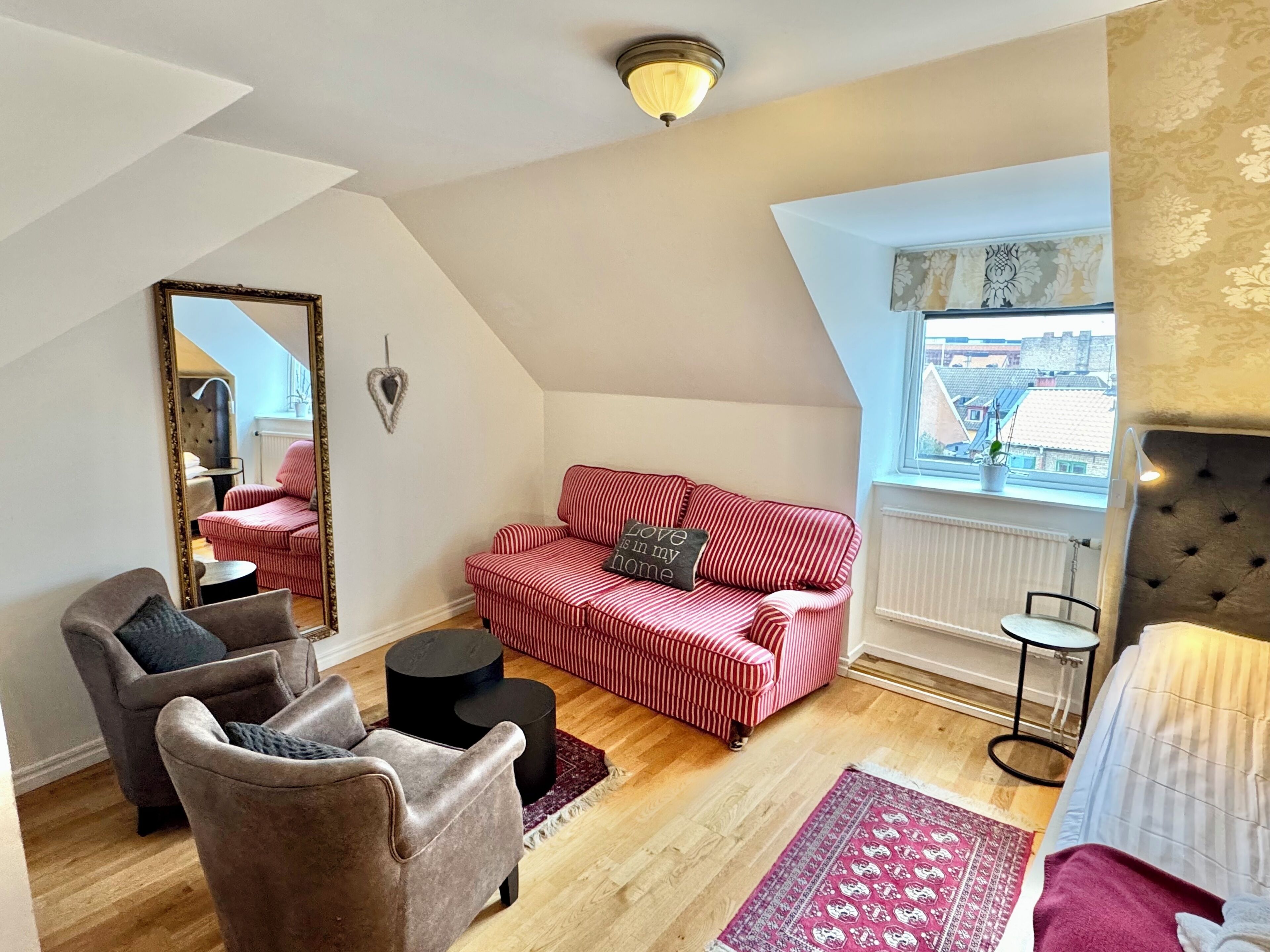 A loft-style room with a red-striped sofa, a brown armchair, a small round table, a mirror, and a window with a view of buildings.