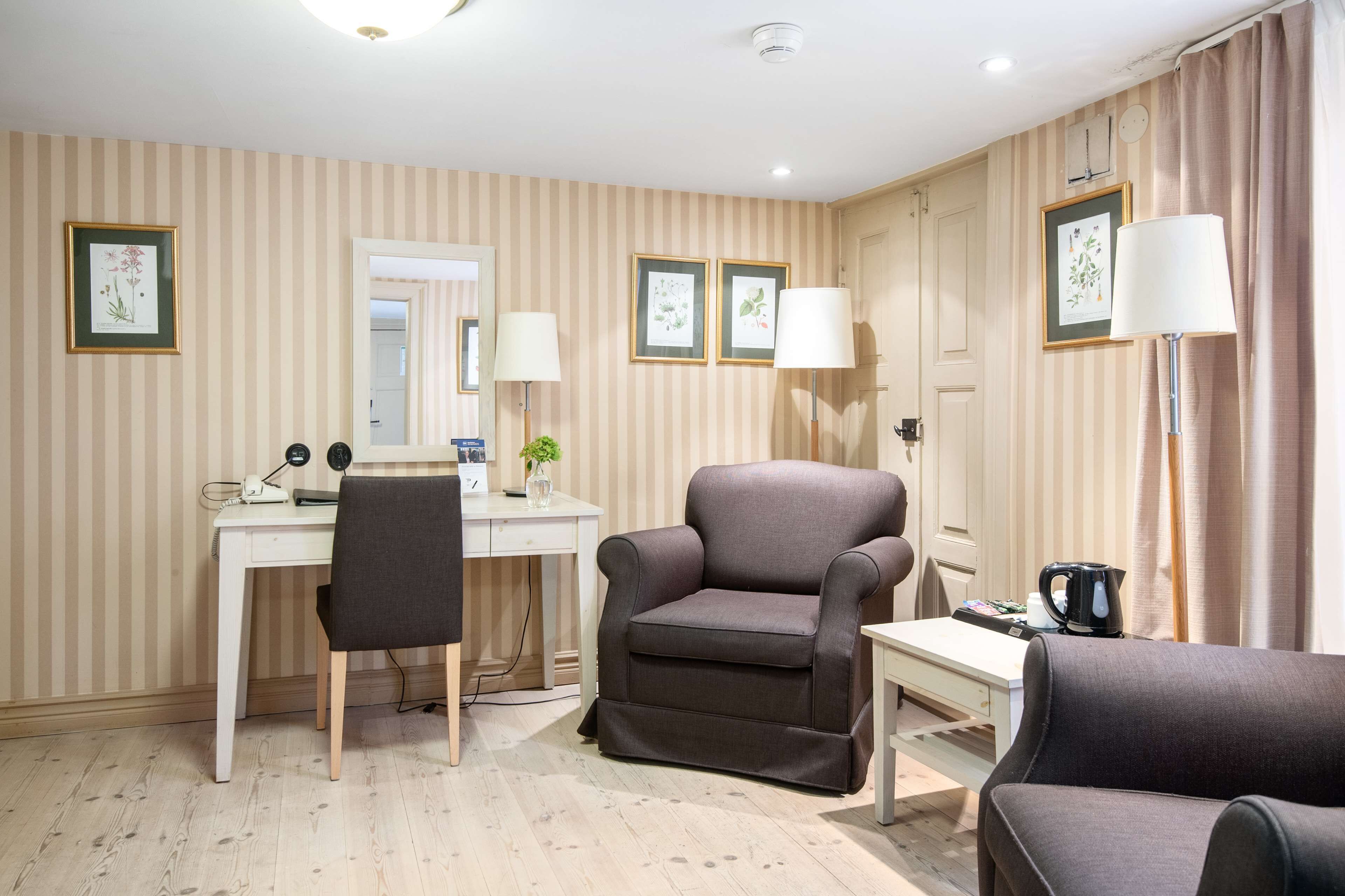 A hotel room with striped wallpaper, a desk with a chair, a sofa, and two lamps.