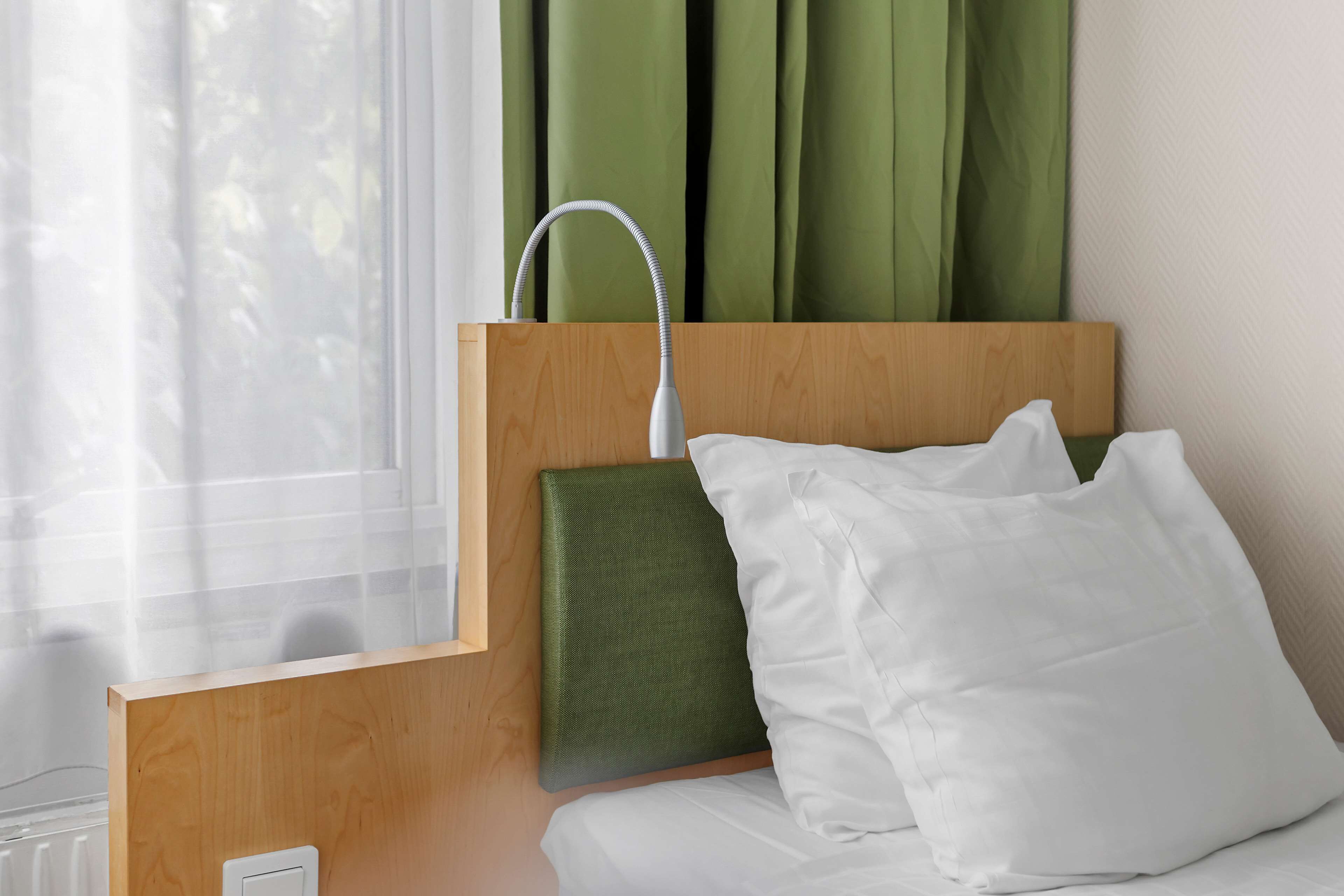 A hotel room with a wooden headboard, a green upholstered headrest, white bedding, and a window with sheer curtains.
