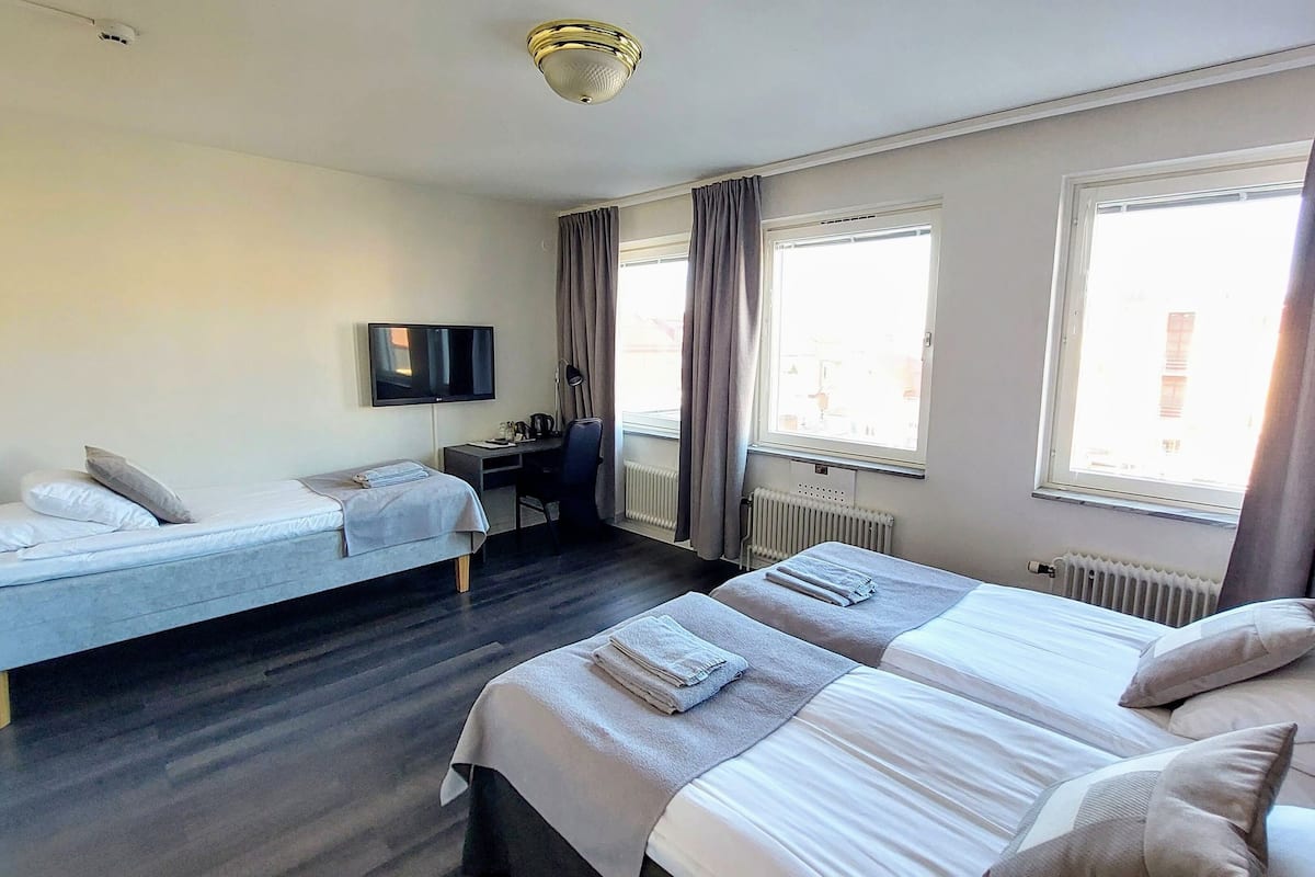 A hotel room with two beds, a desk, a chair, a TV, and large windows with curtains.