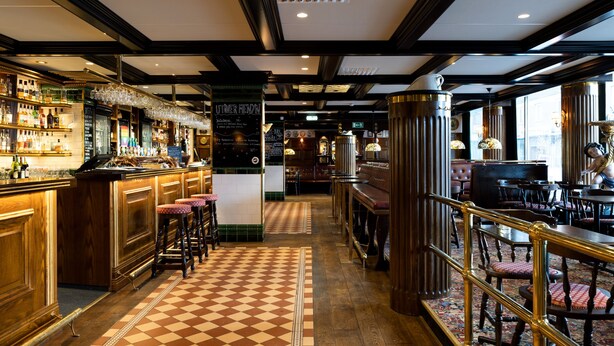 A bar area with a checkered floor, wooden bar counter, stools, and a menu board. - Hotel Bishops Arms Mora (Mora)