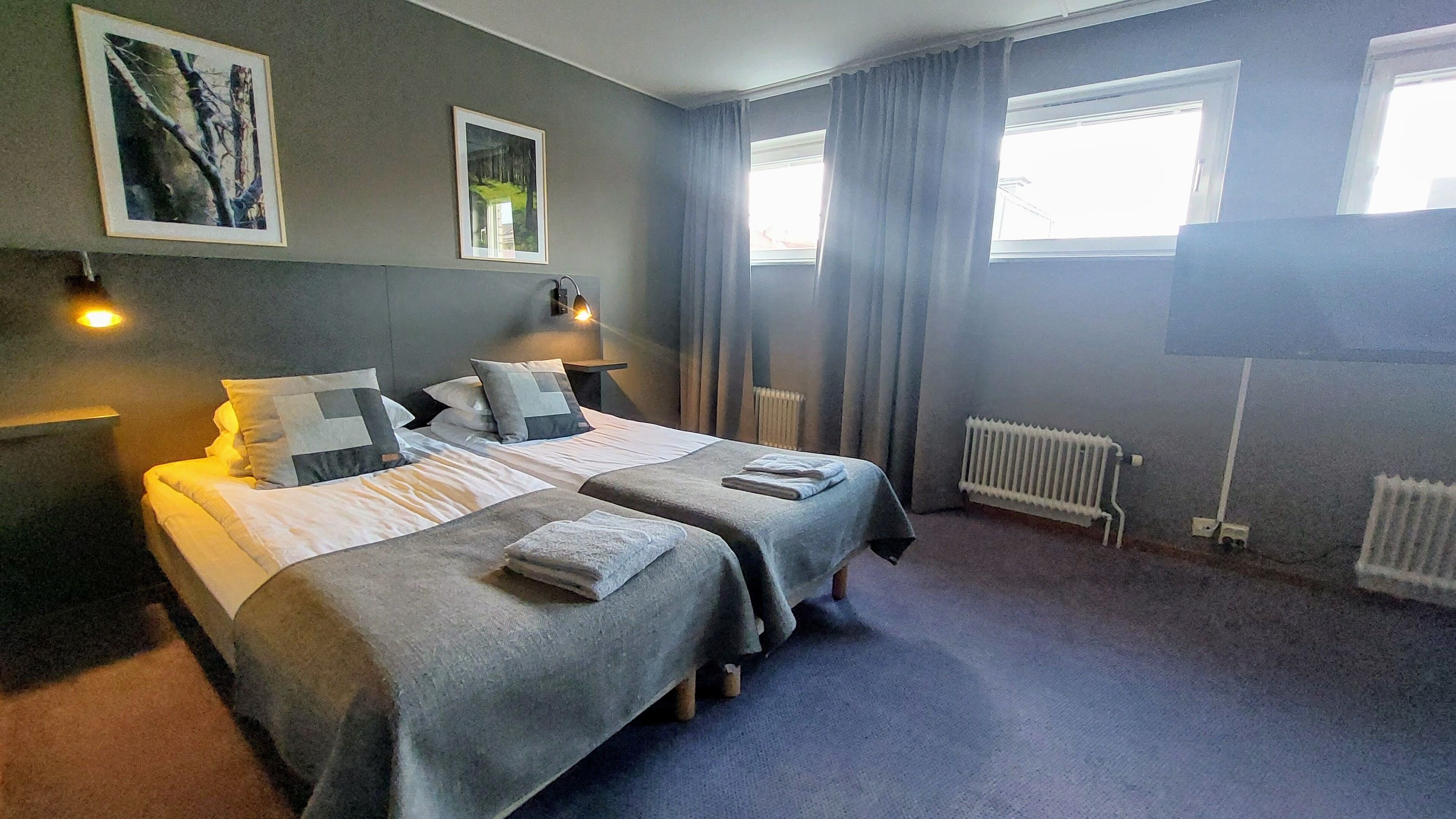 A hotel room with two beds, a radiator, a window with curtains, and wall-mounted lights.