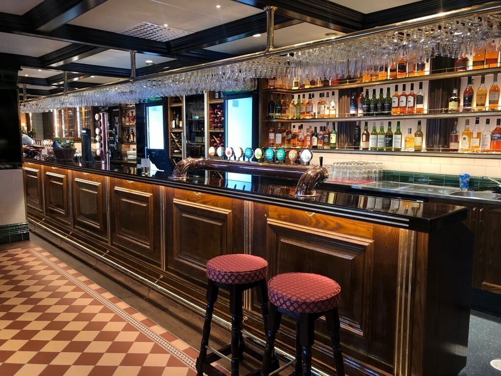 Zona de bar con piso a cuadros, barra de madera, taburetes y una variedad de botellas y vasos en estanterías.
