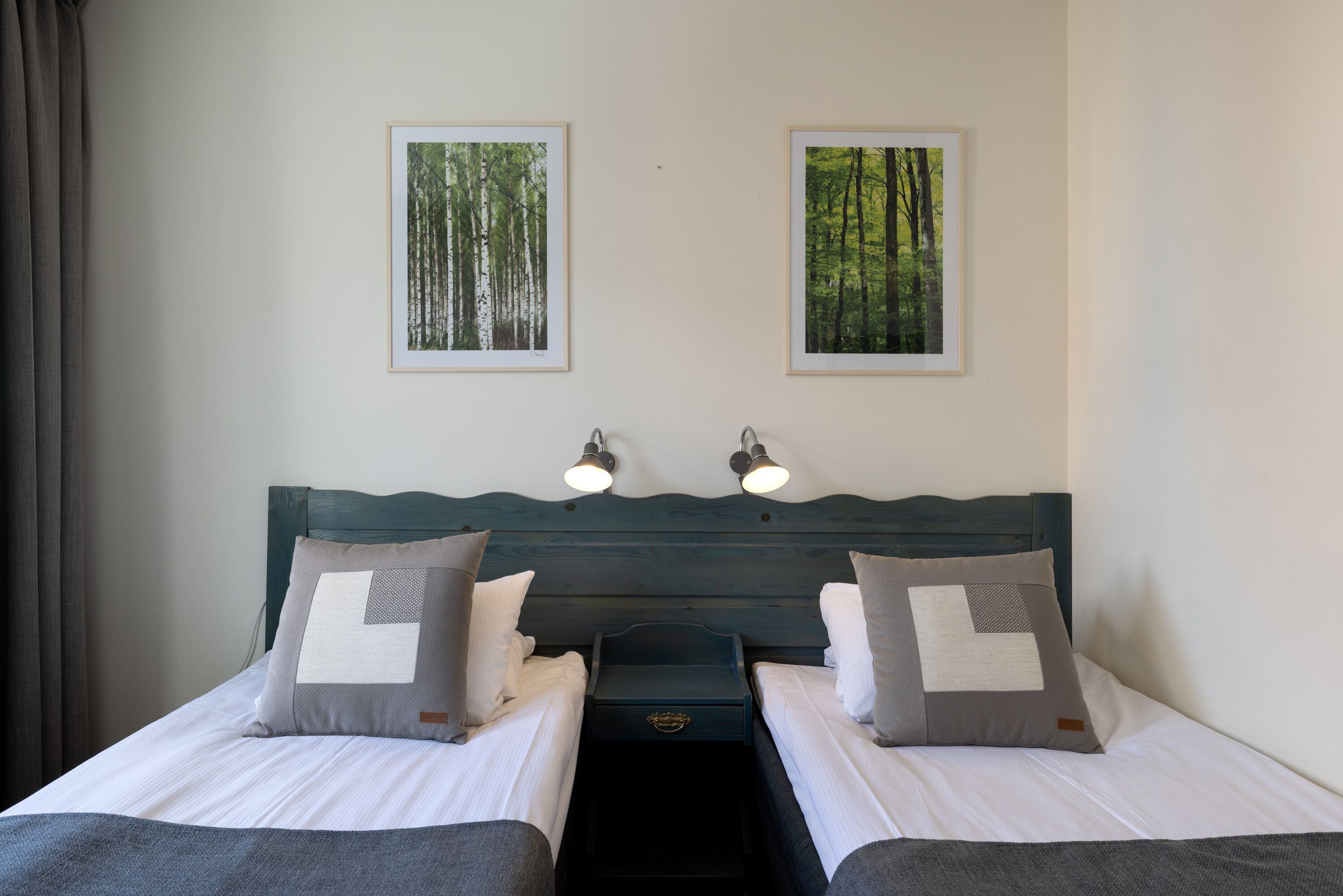 A double bed with two pillows and a nightstand, a headboard with wall-mounted lights, and two framed forest pictures.