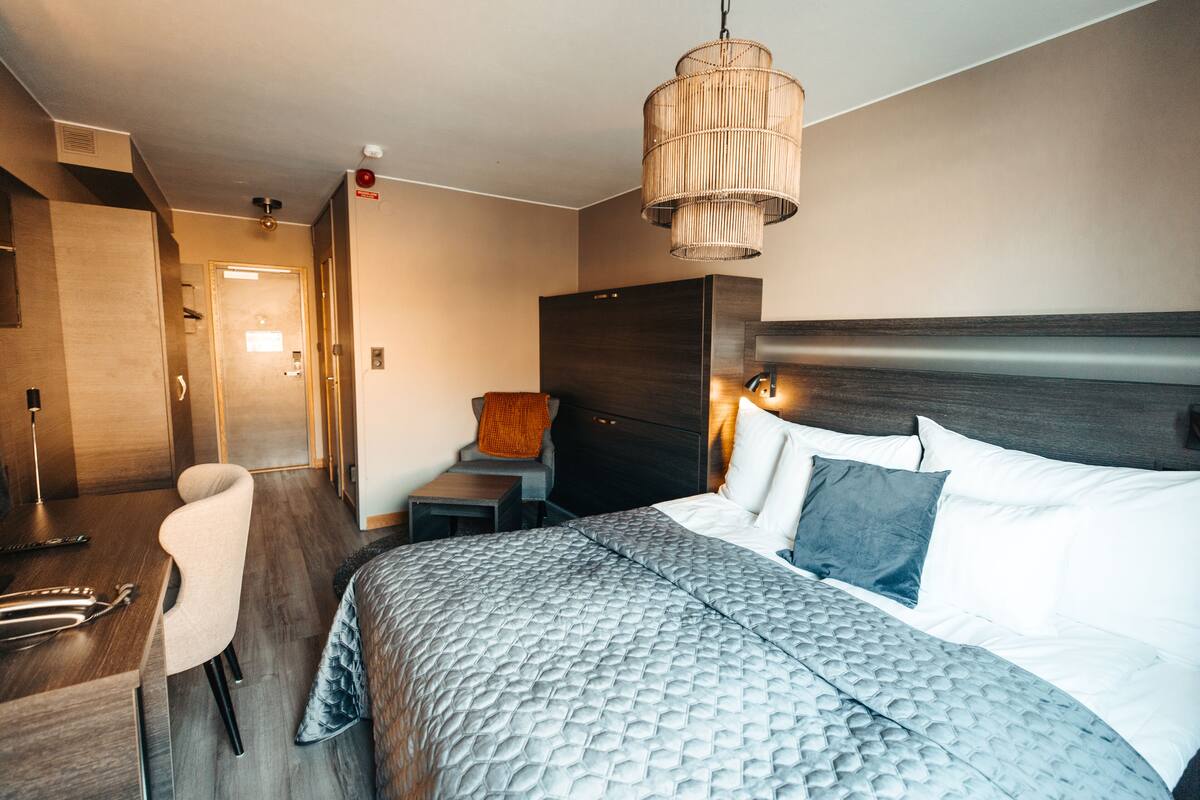 A modern hotel room with a large bed, a bedside table, a chair, and a hanging light fixture.
