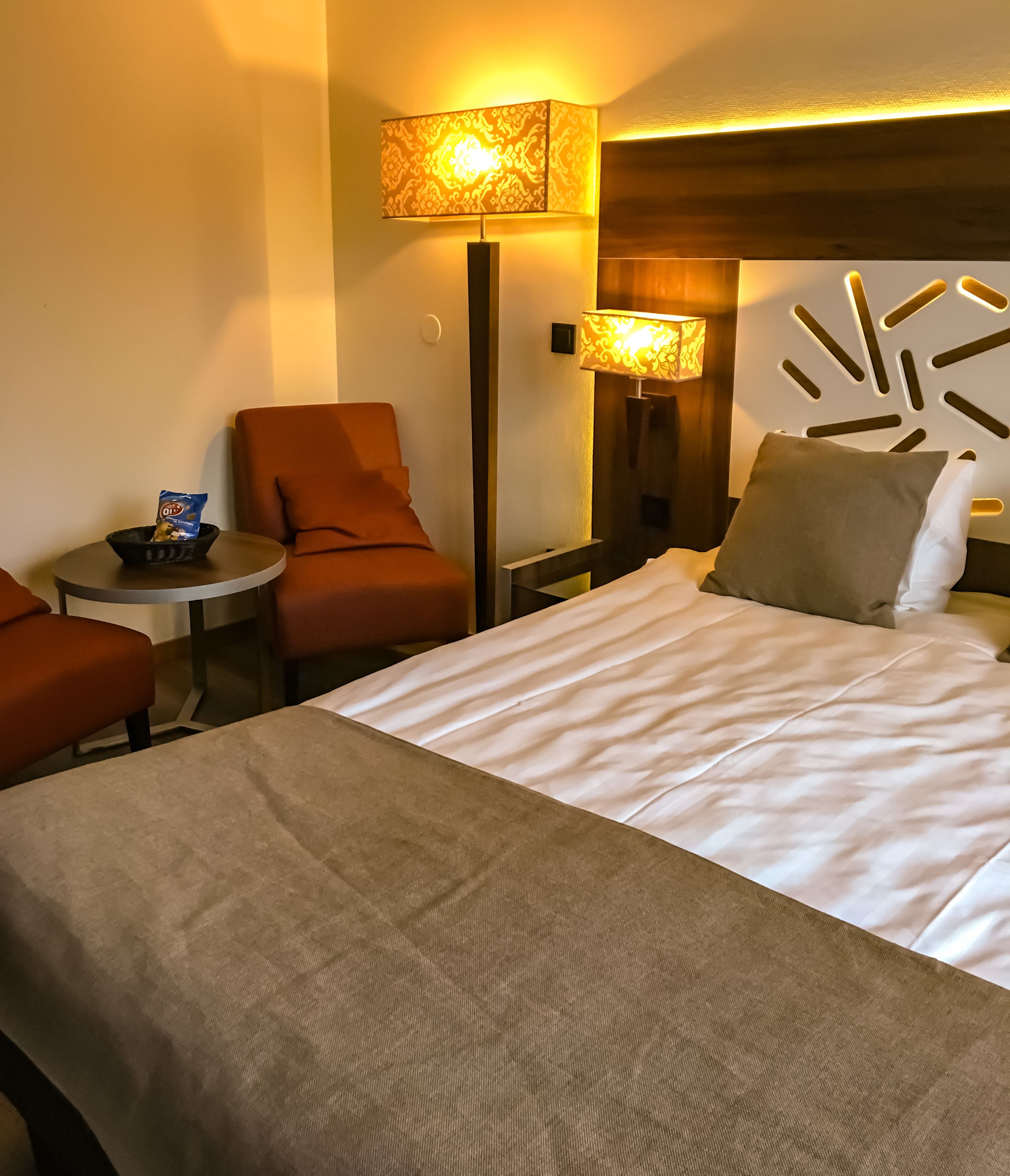 A hotel room with a bed, two armchairs, a small table with snacks, and two lamps.