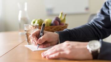 A person writing at a table with a pen, a glass of water, and a basket of fruits in the background.
