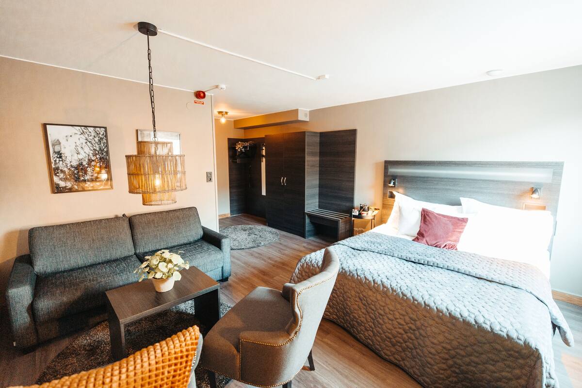 A modern hotel room with a sofa, armchair, and a bed with a quilted cover. There is a small table with a vase of flowers and a hanging light fixture.