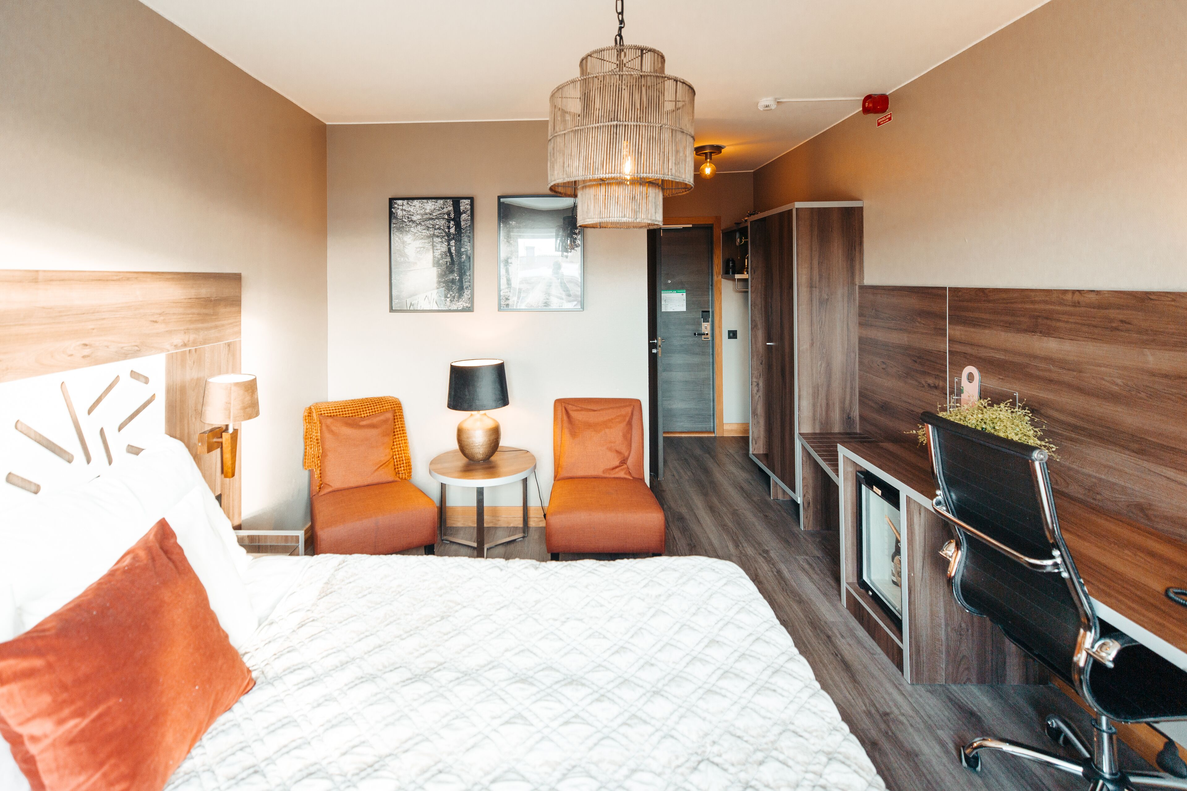 A hotel room with a large bed, two orange chairs, a small table with a lamp, a wooden cabinet, and a TV.