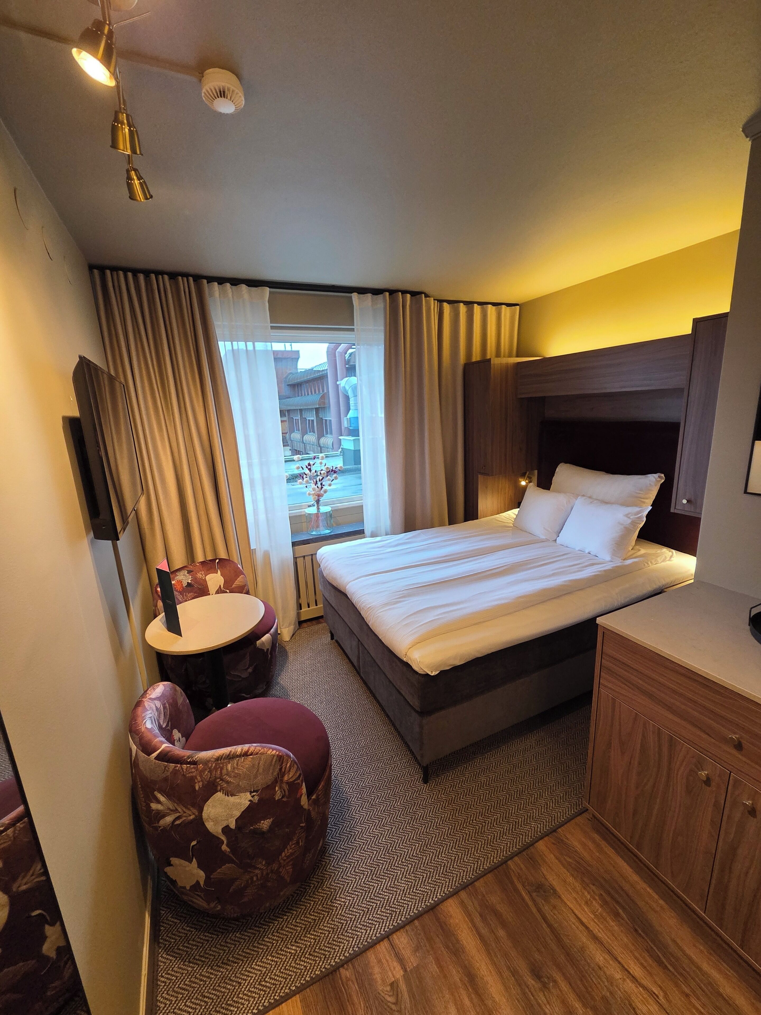 Habitación de hotel con cama, escritorio, silla, ventana con cortinas e iluminación en el techo.