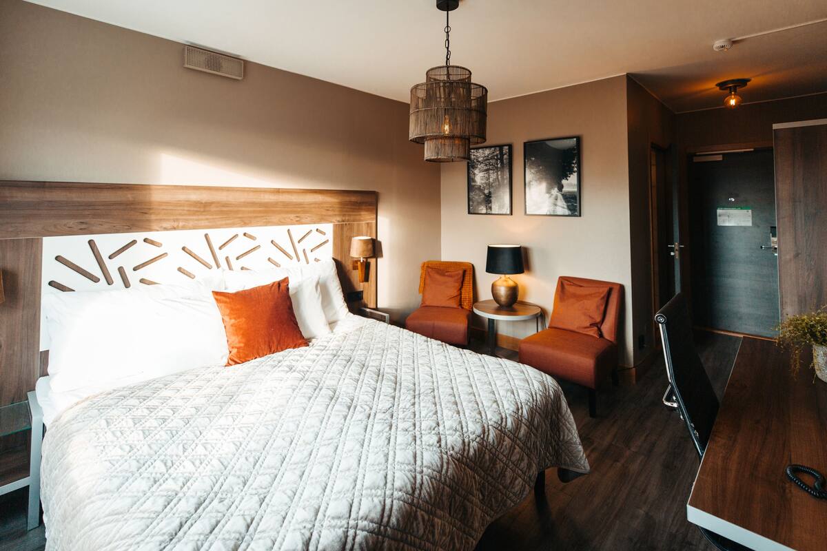 A hotel room with a large bed, a desk, two chairs, a small table, and a hanging light fixture.