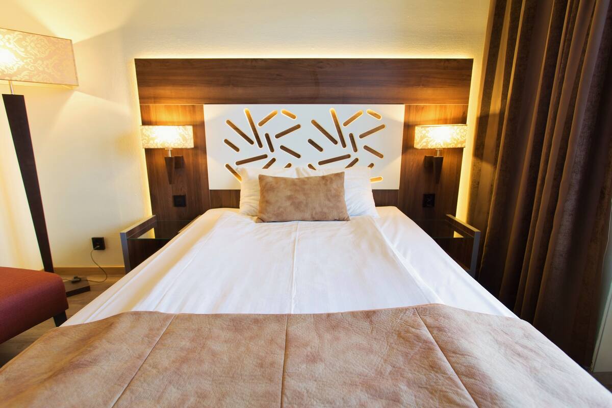 A hotel room with a large bed, two bedside lamps, a wooden headboard, and a red bench.