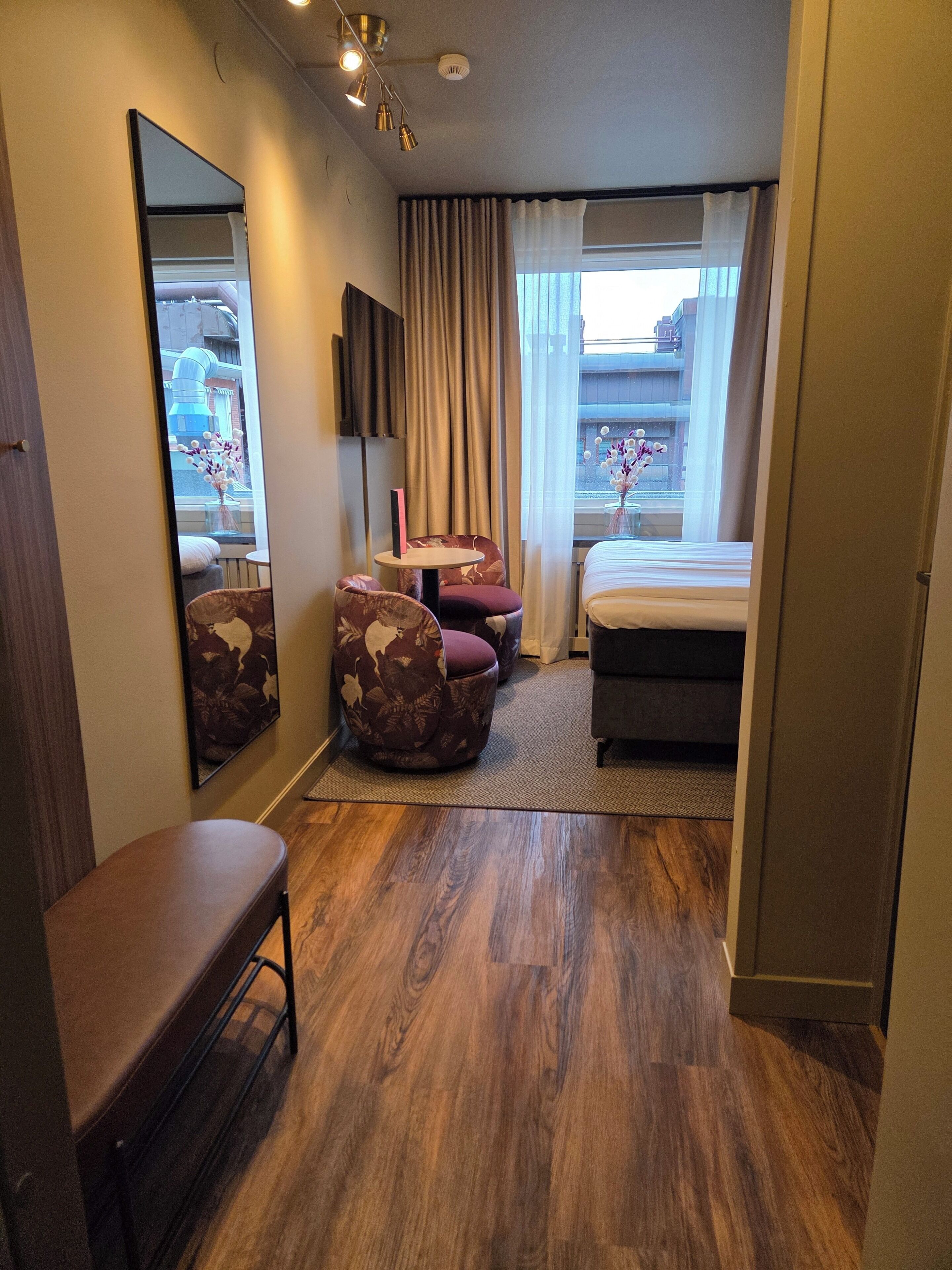 Une chambre d’hôtel équipée d’un lit, d’un bureau, d’une chaise, d’un canapé, d’un miroir et d’une fenêtre avec des rideaux.