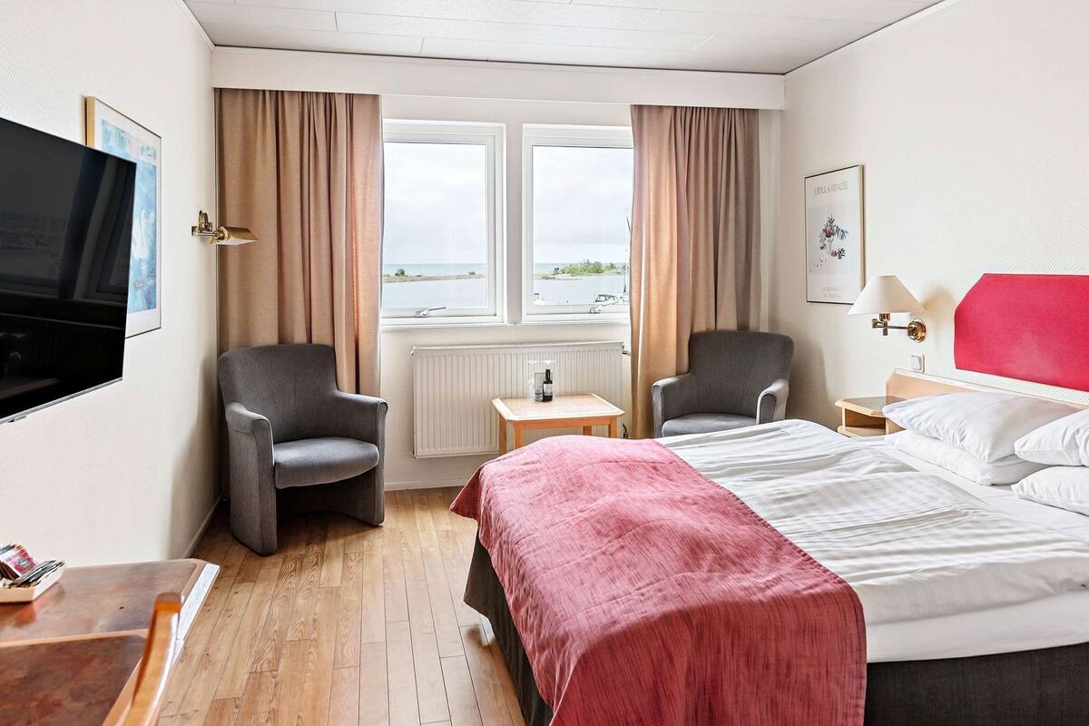 Une chambre d’hôtel avec deux chaises, un lit, une télévision, une fenêtre donnant sur un beau paysage et une tête de lit rouge.