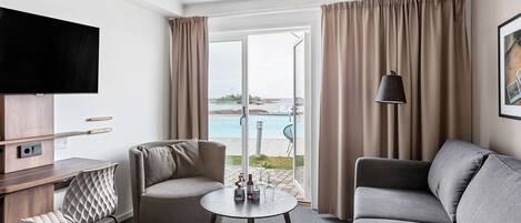 A modern hotel room with a flat-screen TV, a sofa, a chair, a small round table, and a balcony with a pool view.