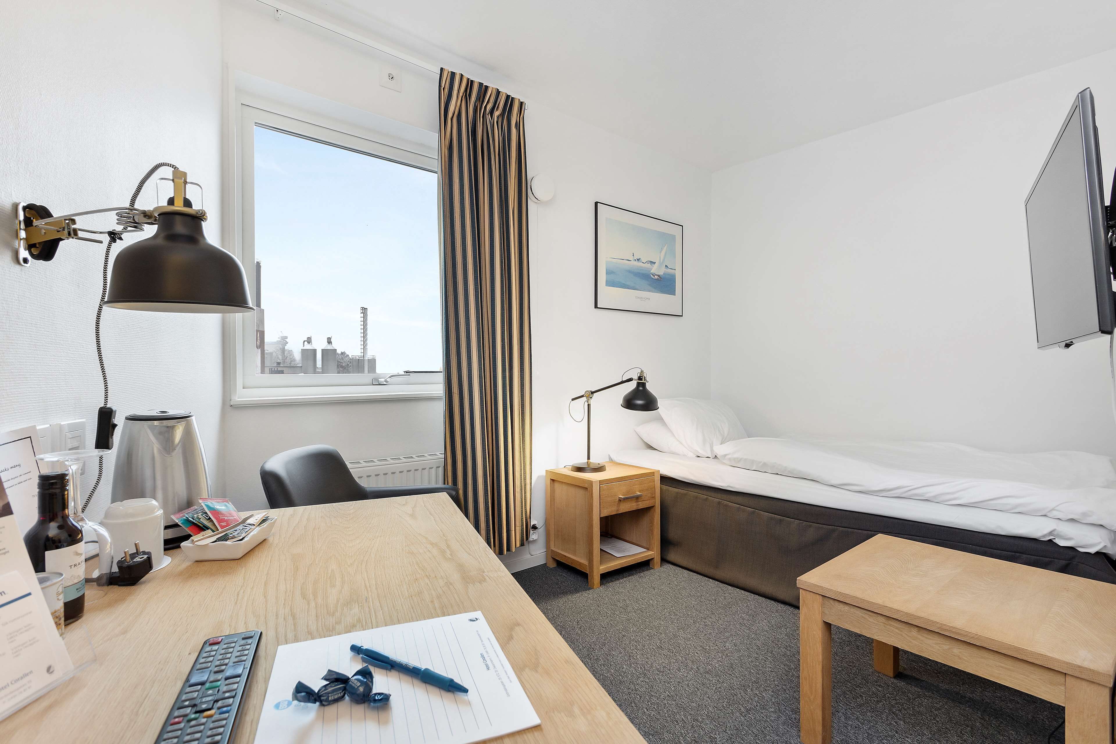 Habitación de hotel con cama, escritorio con lámpara, silla, una mesita, ventana con cortinas y vista a la ciudad.