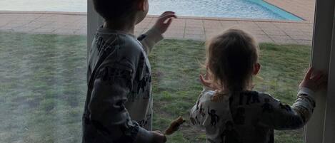 Two children looking out a window at a swimming pool and a distant shoreline.