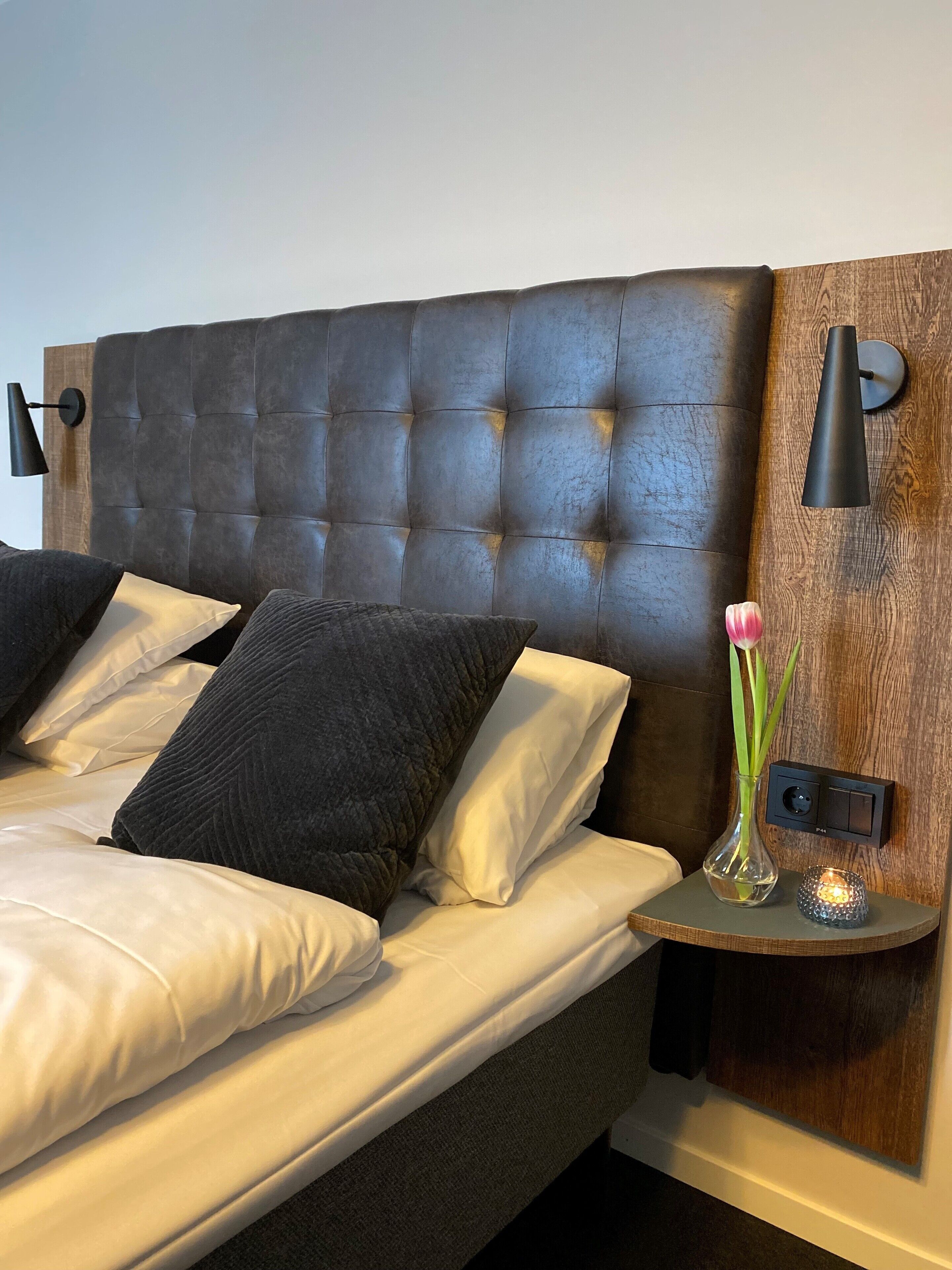 Ein Schlafzimmer mit einem gepolsterten Kopfteil, einem Nachttisch mit Vase und Kerze sowie einer Wandleuchte.