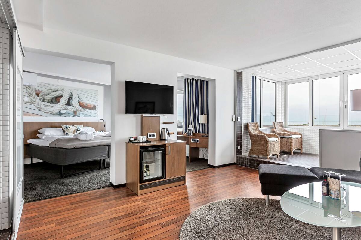 Une chambre d’hôtel moderne équipée d’un lit, d’un minibar, d’une télévision à écran plat, d’un coin salon avec une table en verre et offrant une vue sur la mer grâce à de grandes fenêtres.