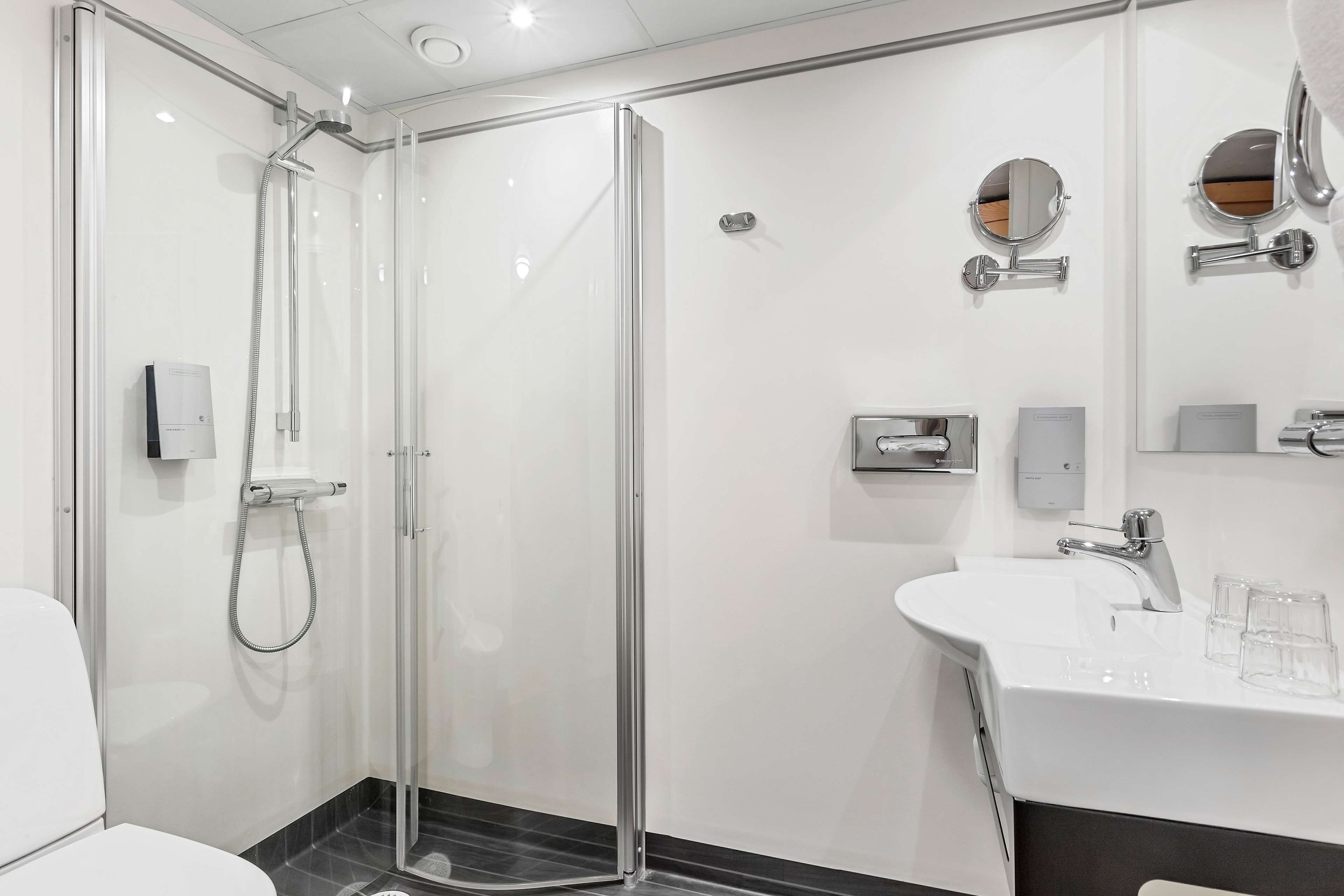 A modern bathroom with a glass-enclosed shower, a white sink with two glasses, and a mirror with a round frame.