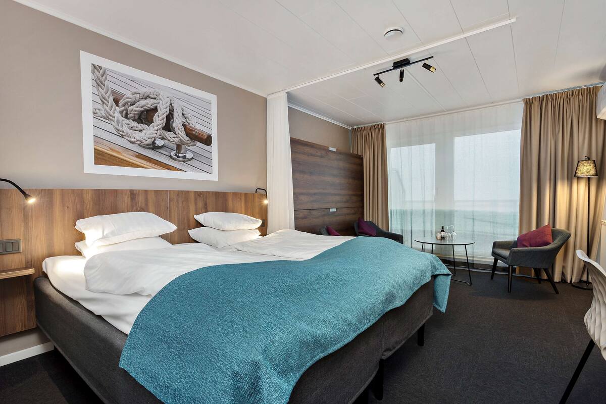 Une chambre d’hôtel avec un lit, un bureau, une chaise, une fenêtre avec des rideaux et un tableau au mur.