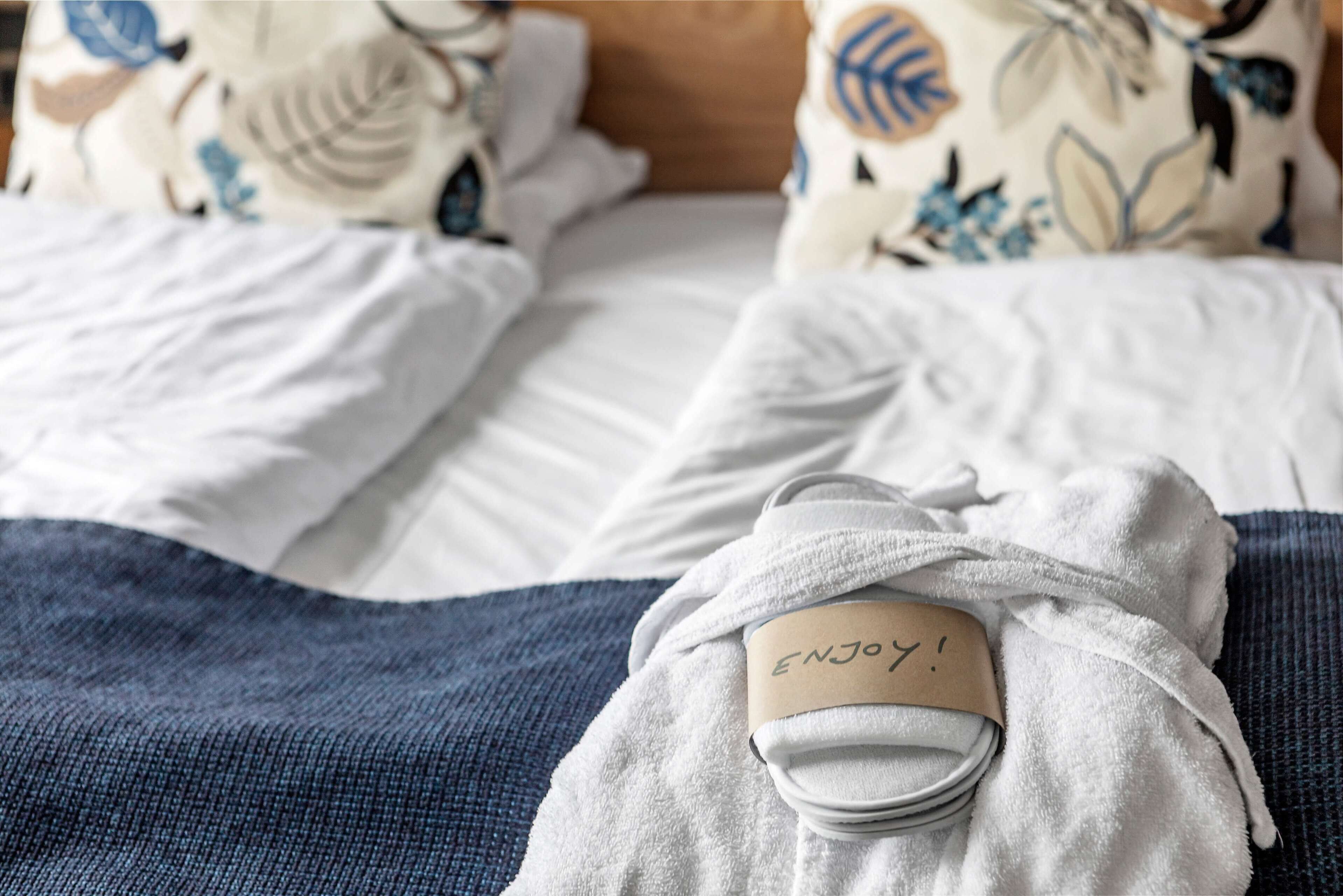 Una cama bien tendida con una toalla blanca y una etiqueta que dice "ENJOY!" sobre un cojín de estampado azul y blanco.