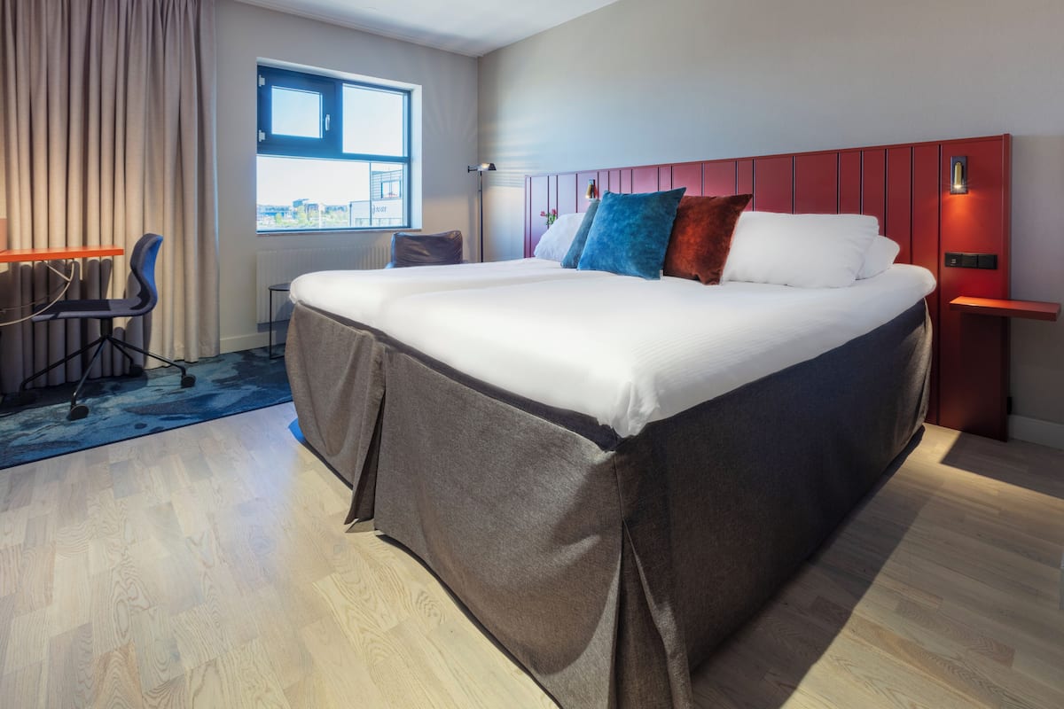 Ein modernes Hotelzimmer mit einem großen Bett, einem Schreibtisch mit Stuhl, einem Fenster mit Aussicht und einer roten Akzentwand.