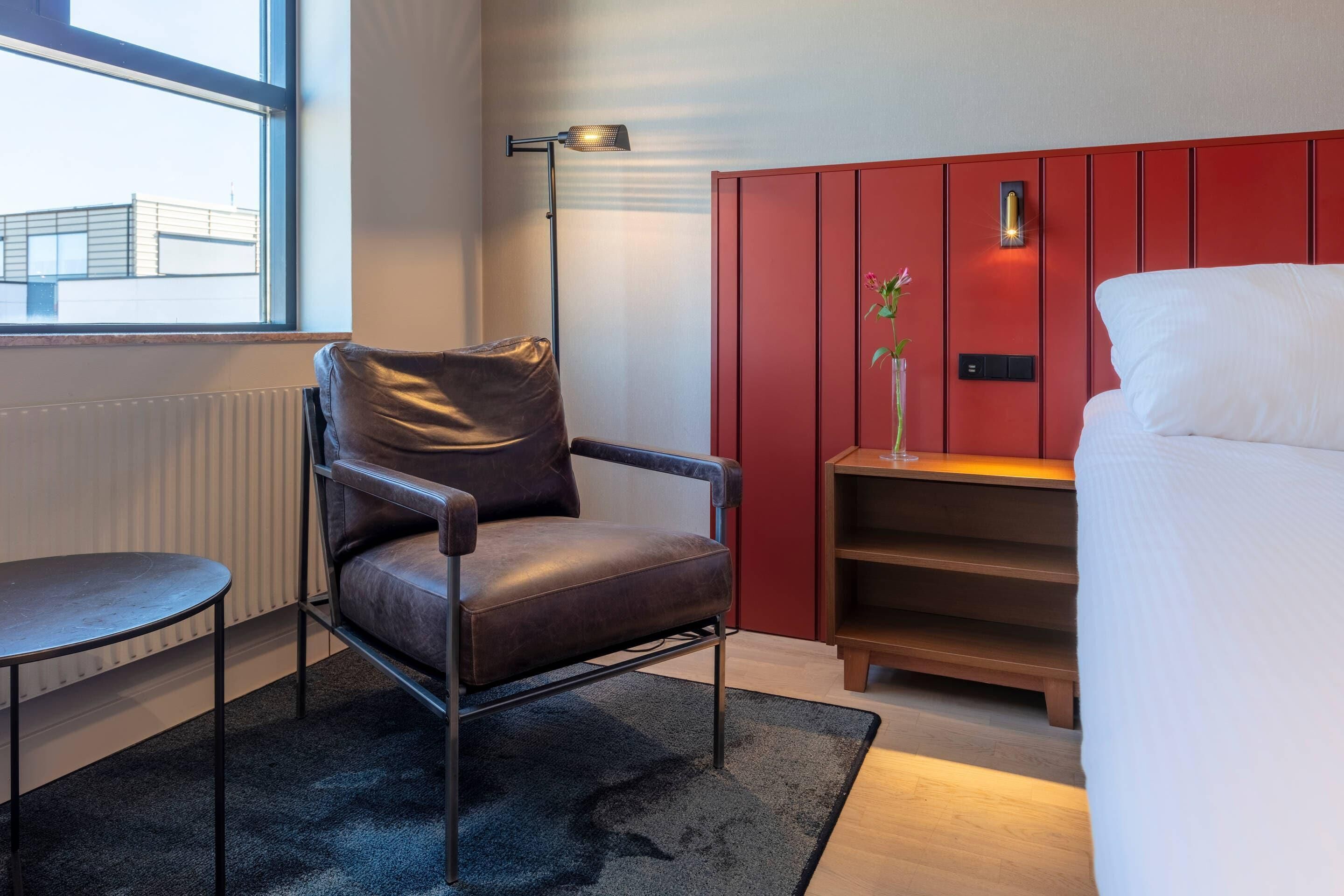 Una habitación de hotel moderna con una butaca de cuero, una mesita, una pared de acento roja, una mesita de noche con un jarrón y una cama.