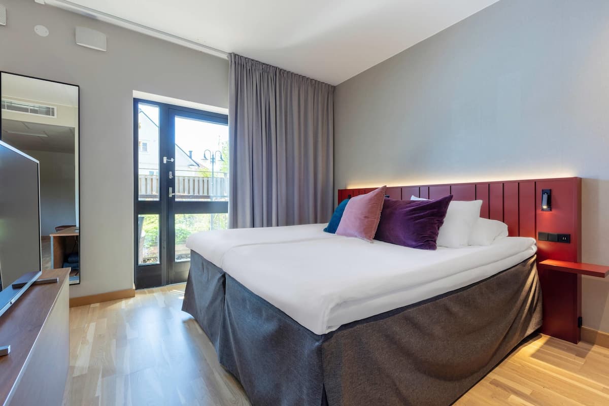 Ein modernes Hotelzimmer mit einem großen Bett, einem Nachttisch, einem Fernseher und einem Fenster mit Vorhängen.