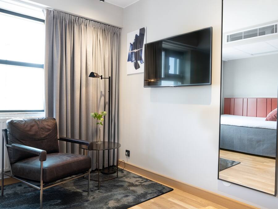 Habitació moderna amb una cadira de pell, una taula petita amb un ram, un televisor fixat a la paret i un llit amb capçalera vermella.