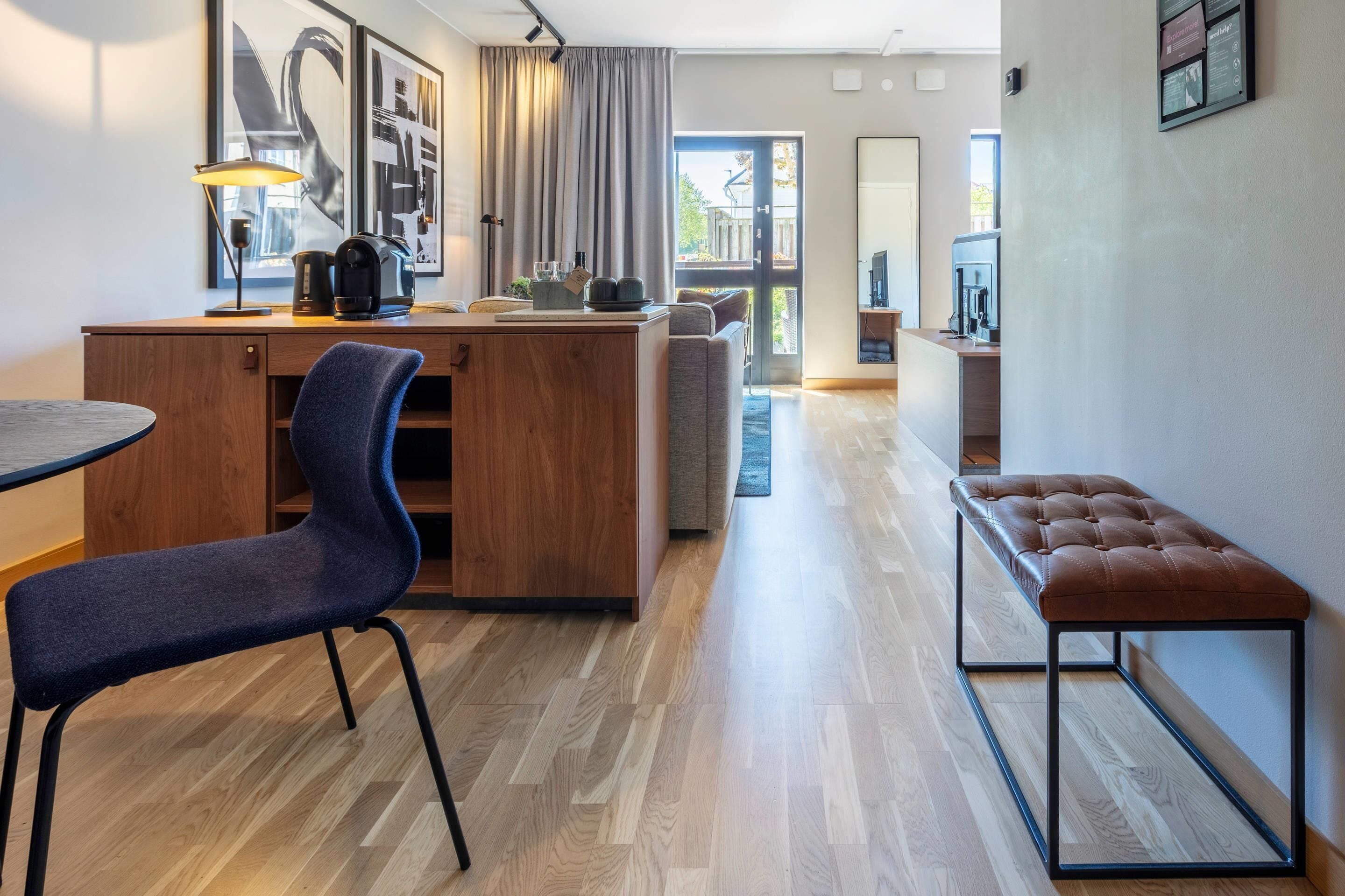 Una habitación de hotel moderna con un escritorio de madera, una silla azul, un banco y un amplio ventanal con vistas al exterior.