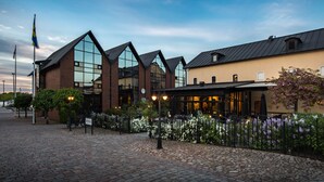 A building with a brick facade and large windows, a paved courtyard, and a restaurant with outdoor seating. - Home Hotel Carlscrona (Karlskrona)