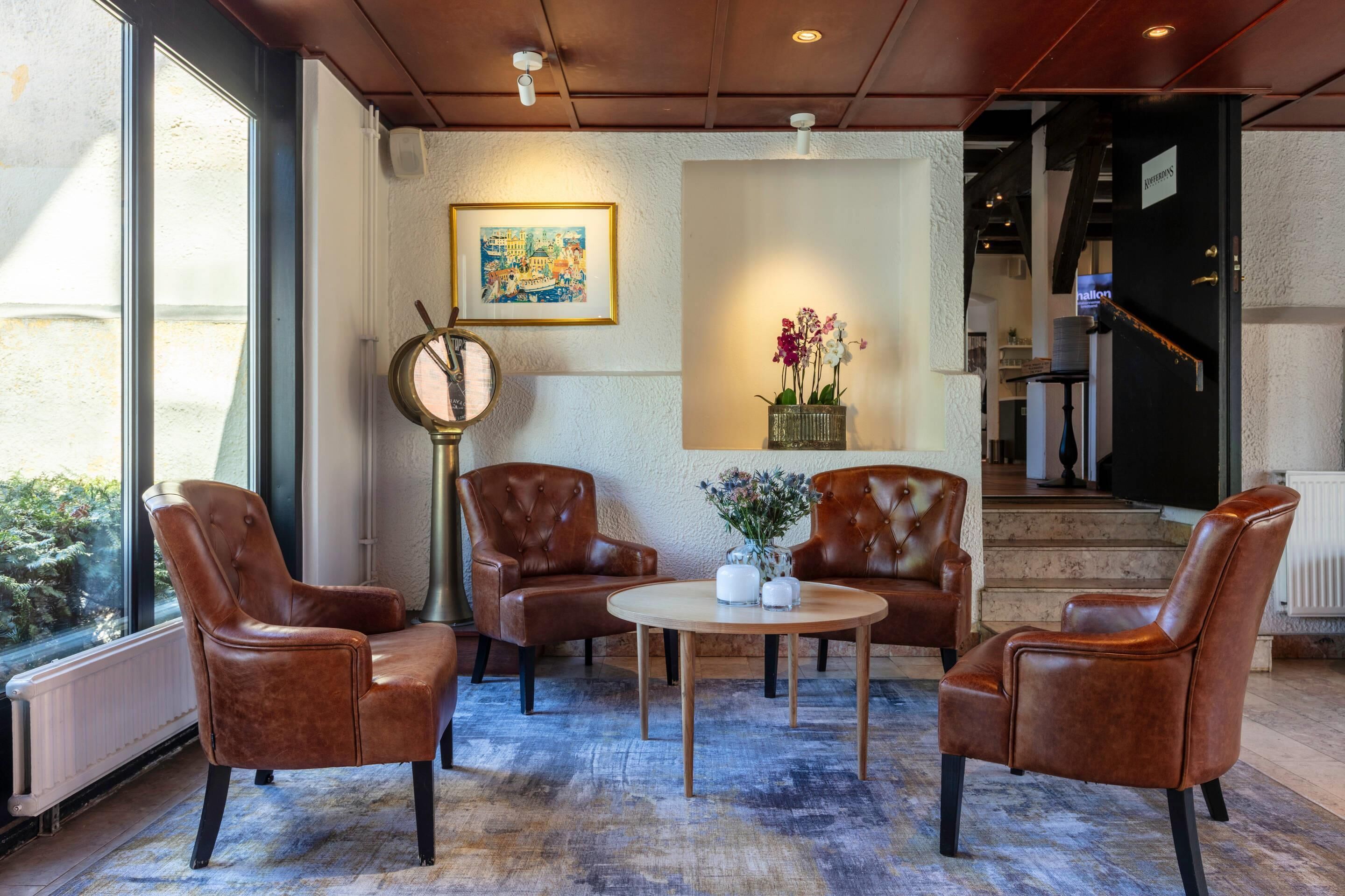 A lobby with leather armchairs, a round table, a framed painting, and a vase with flowers.