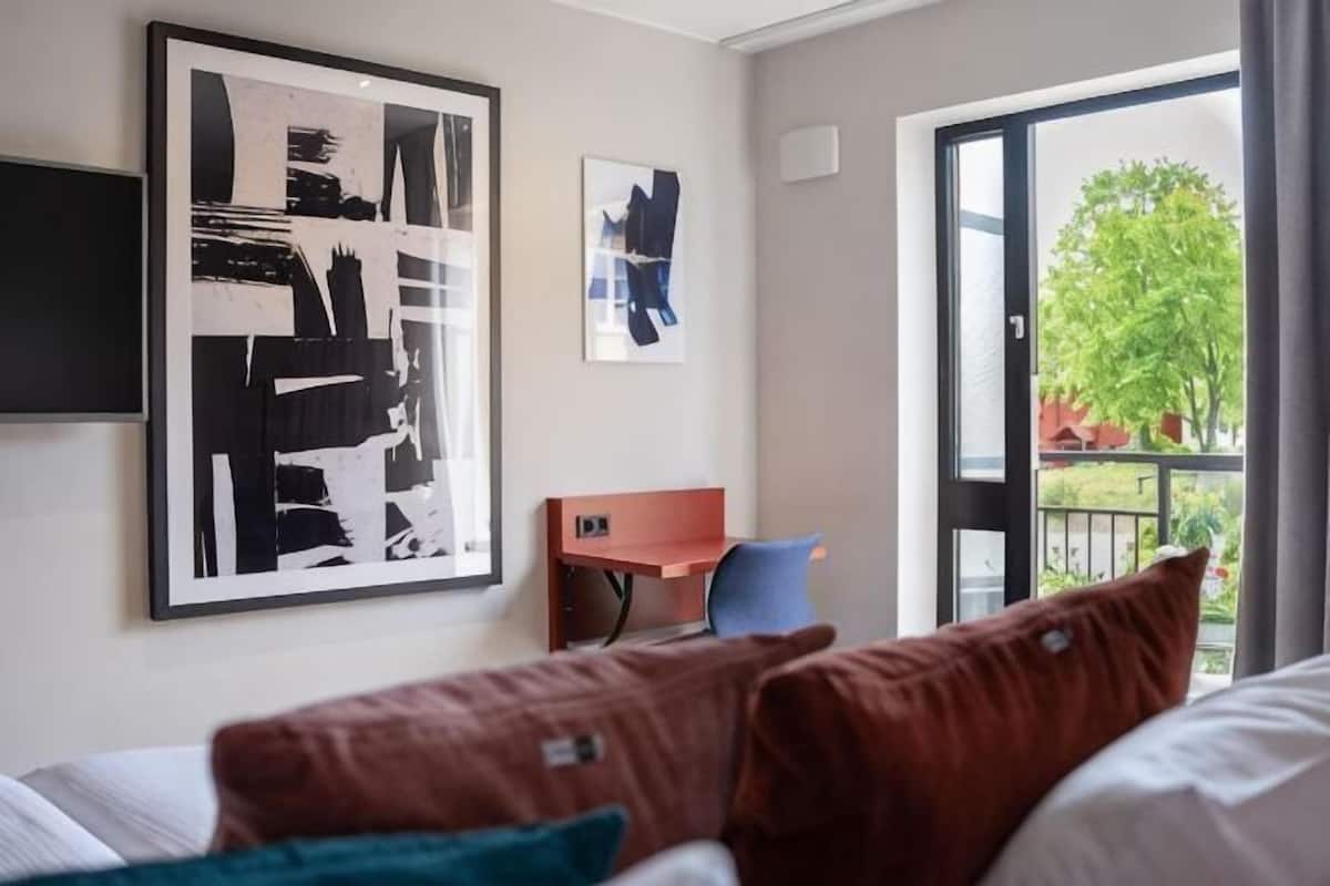 Ein Hotelzimmer mit Bett, Schreibtisch, Stuhl und einem großen Fenster mit Blick auf Grünflächen.