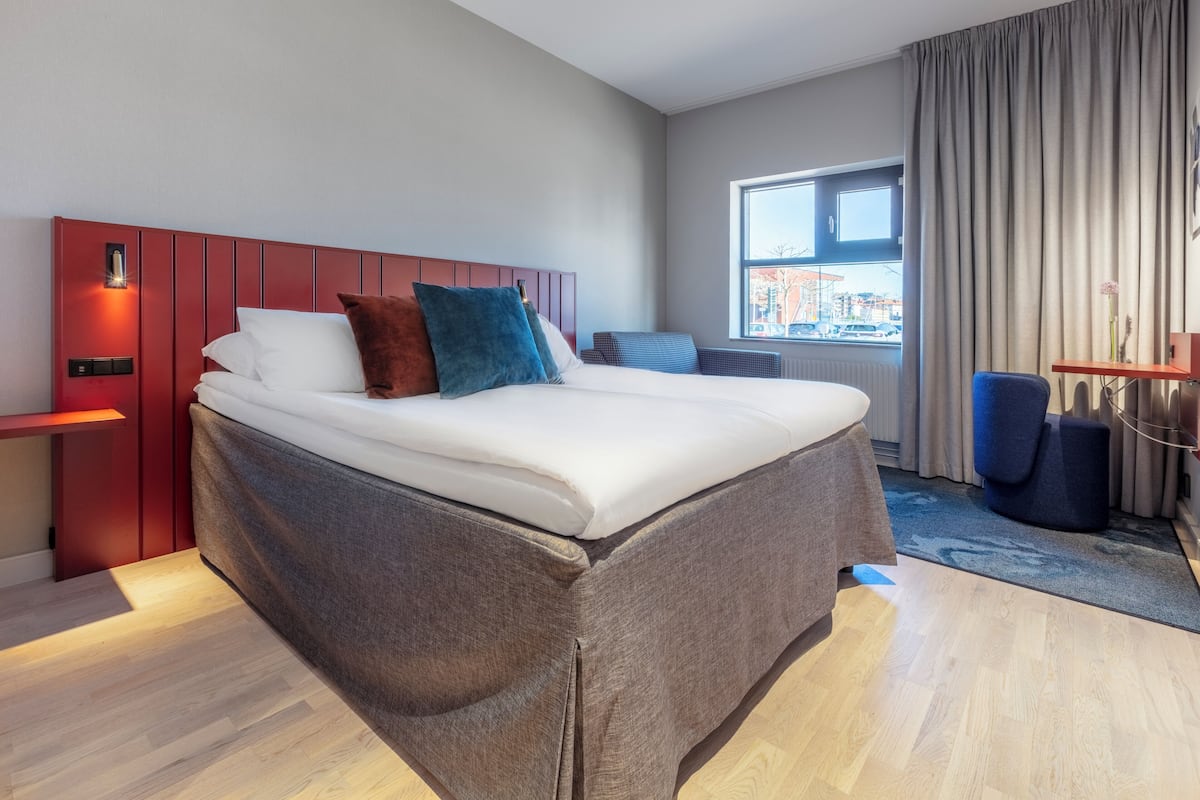 Ein modernes Hotelzimmer mit einem großen Bett, einer roten Kopfteil, einem Fenster mit Aussicht und einem kleinen Schreibtisch mit Stuhl.
