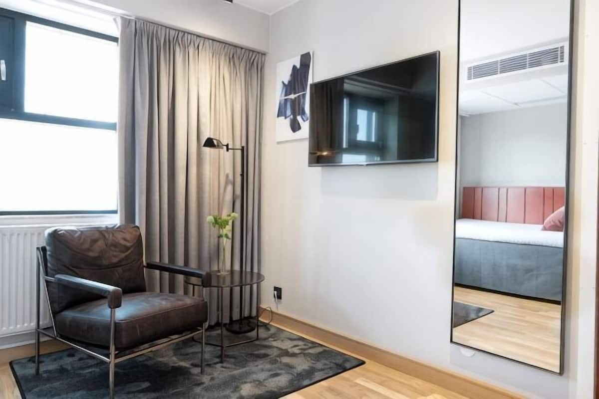 Ein modernes Hotelzimmer mit Ledersessel, kleinem Tisch mit einer Vase, einem Flachbildfernseher und einem großen Spiegel, der ein Bett reflektiert.