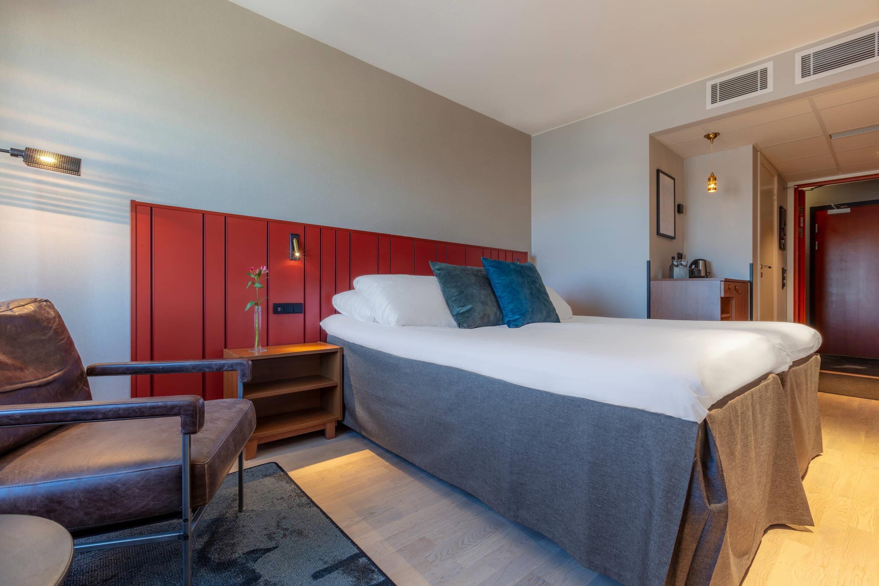 A modern hotel room with a large bed, a red wardrobe, a desk, and a chair.
