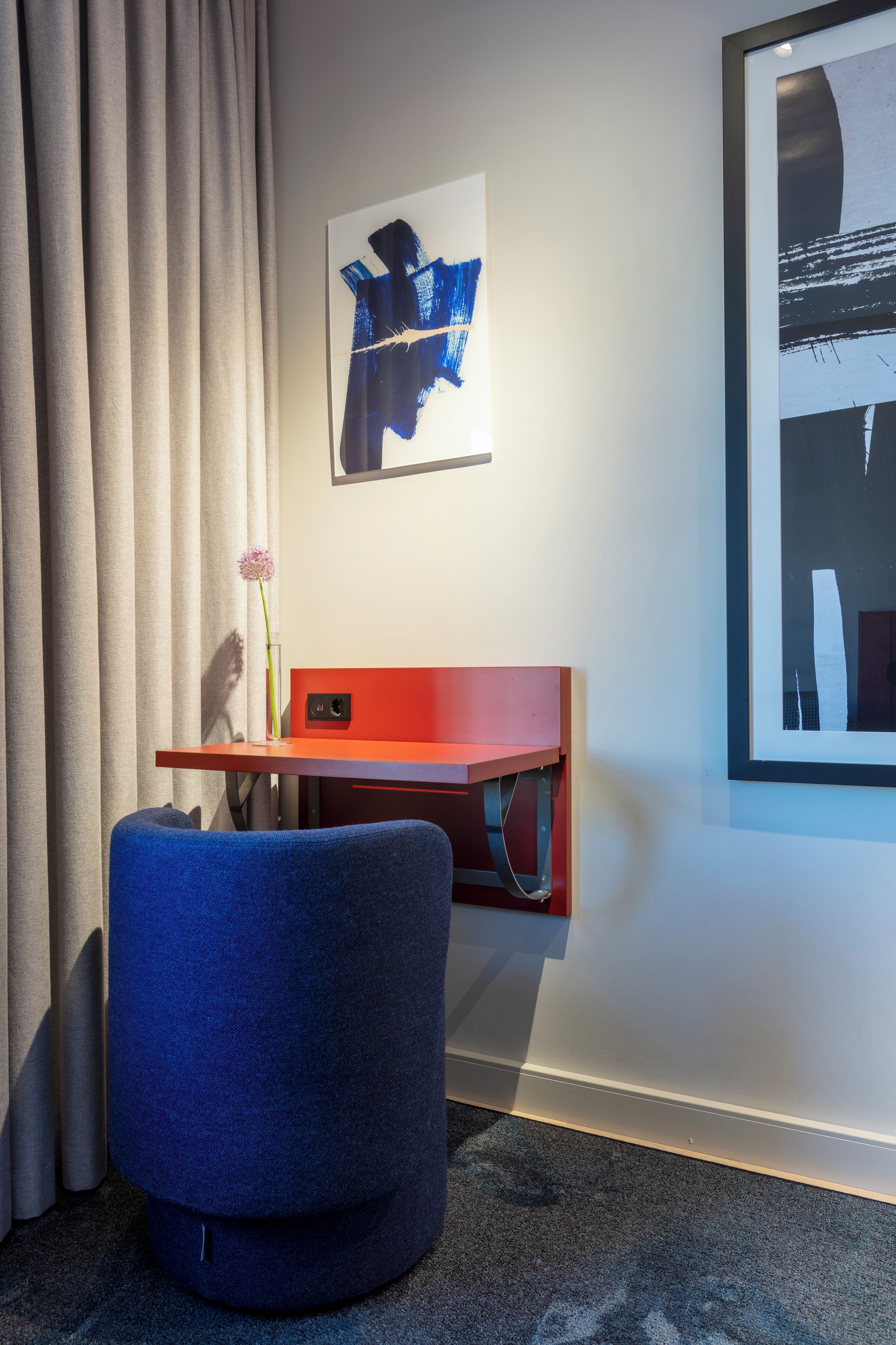 A room with a blue chair, a red desk, a framed abstract painting, and a window with a view.