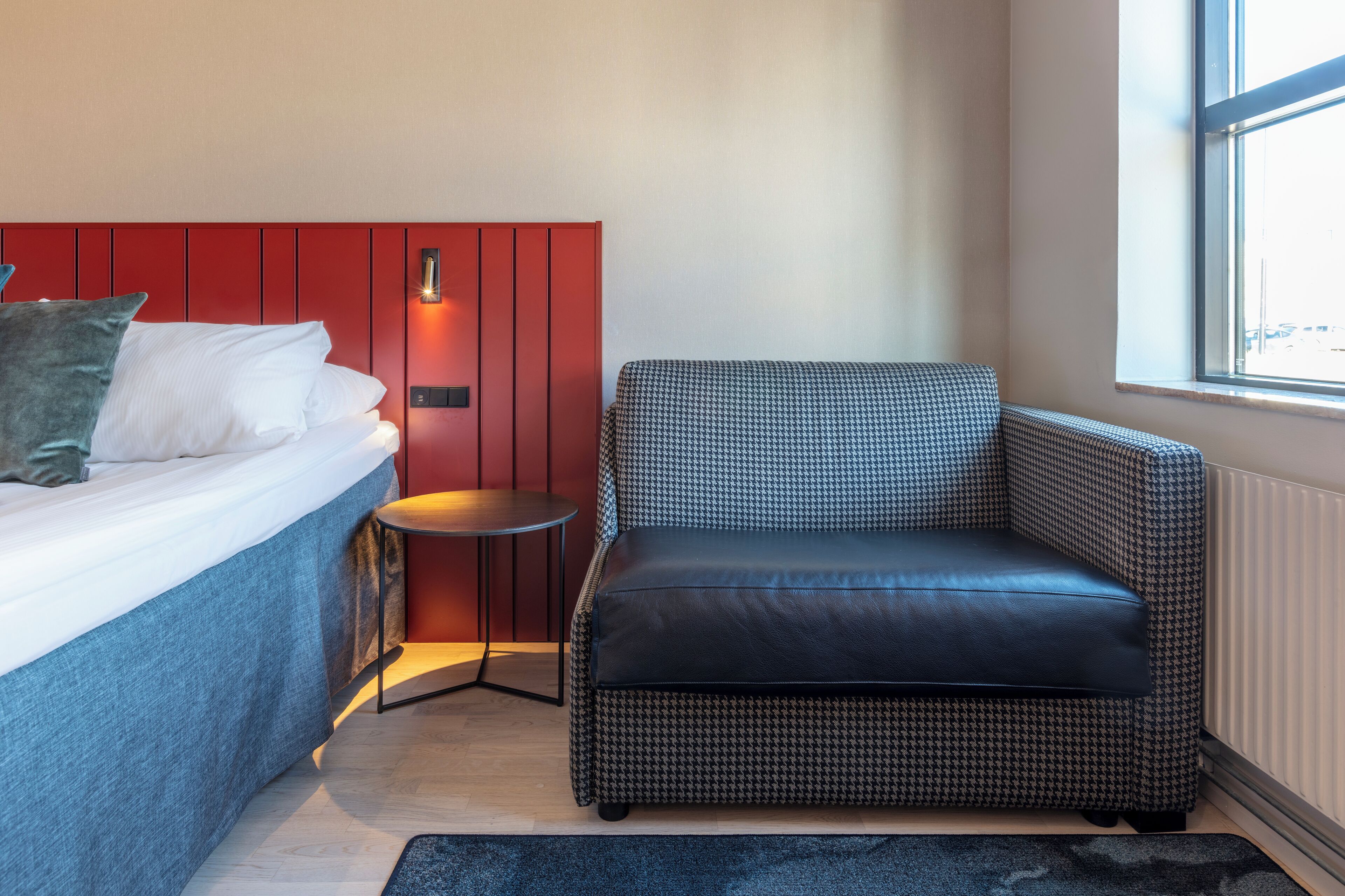 A bedroom with a bed, a red headboard, a houndstooth patterned chair, a small round table, and a window with a view.
