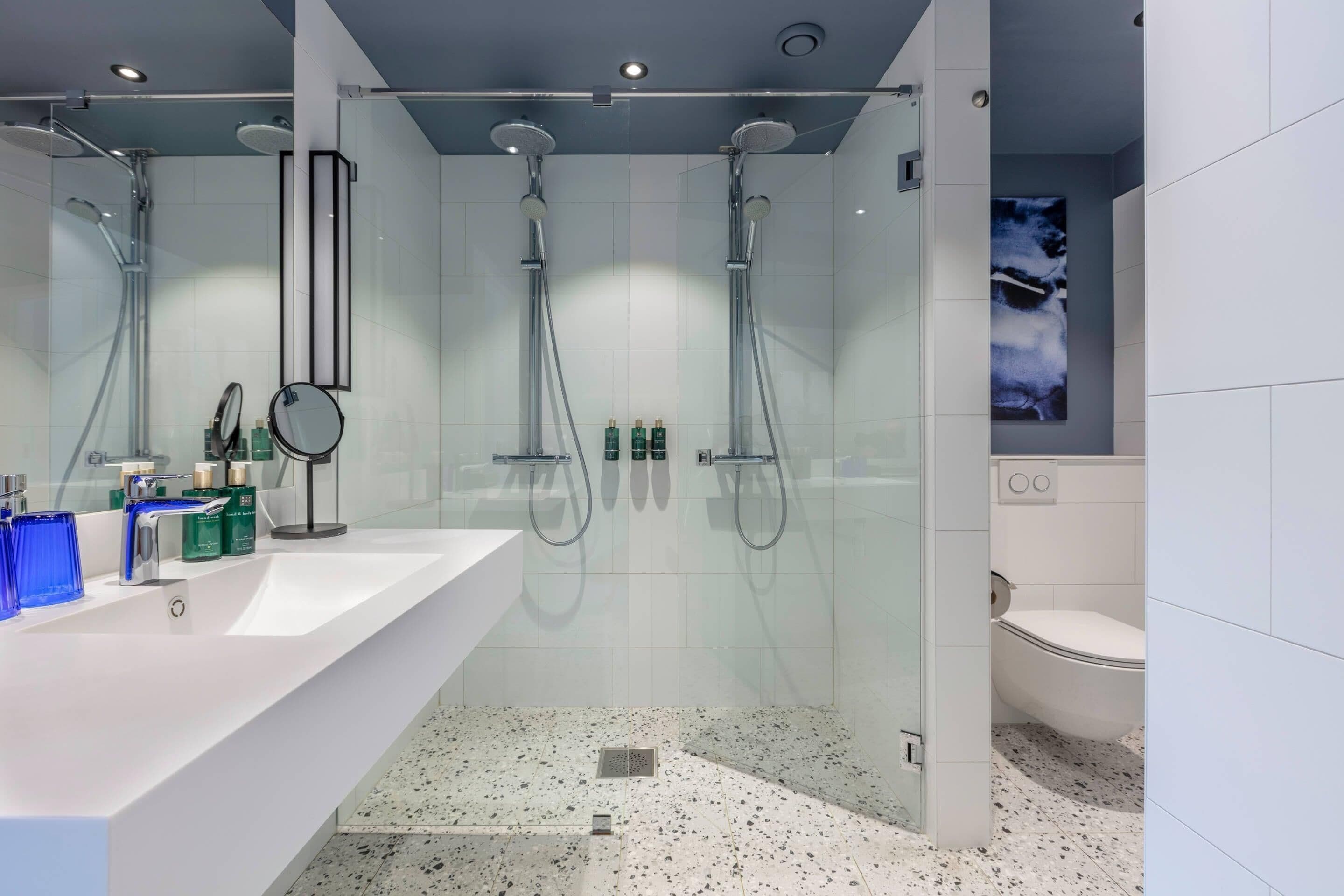 Un baño moderno con un lavamanos grande de color blanco, dos duchas de mano, una cabina de ducha con paredes de vidrio y un inodoro blanco.