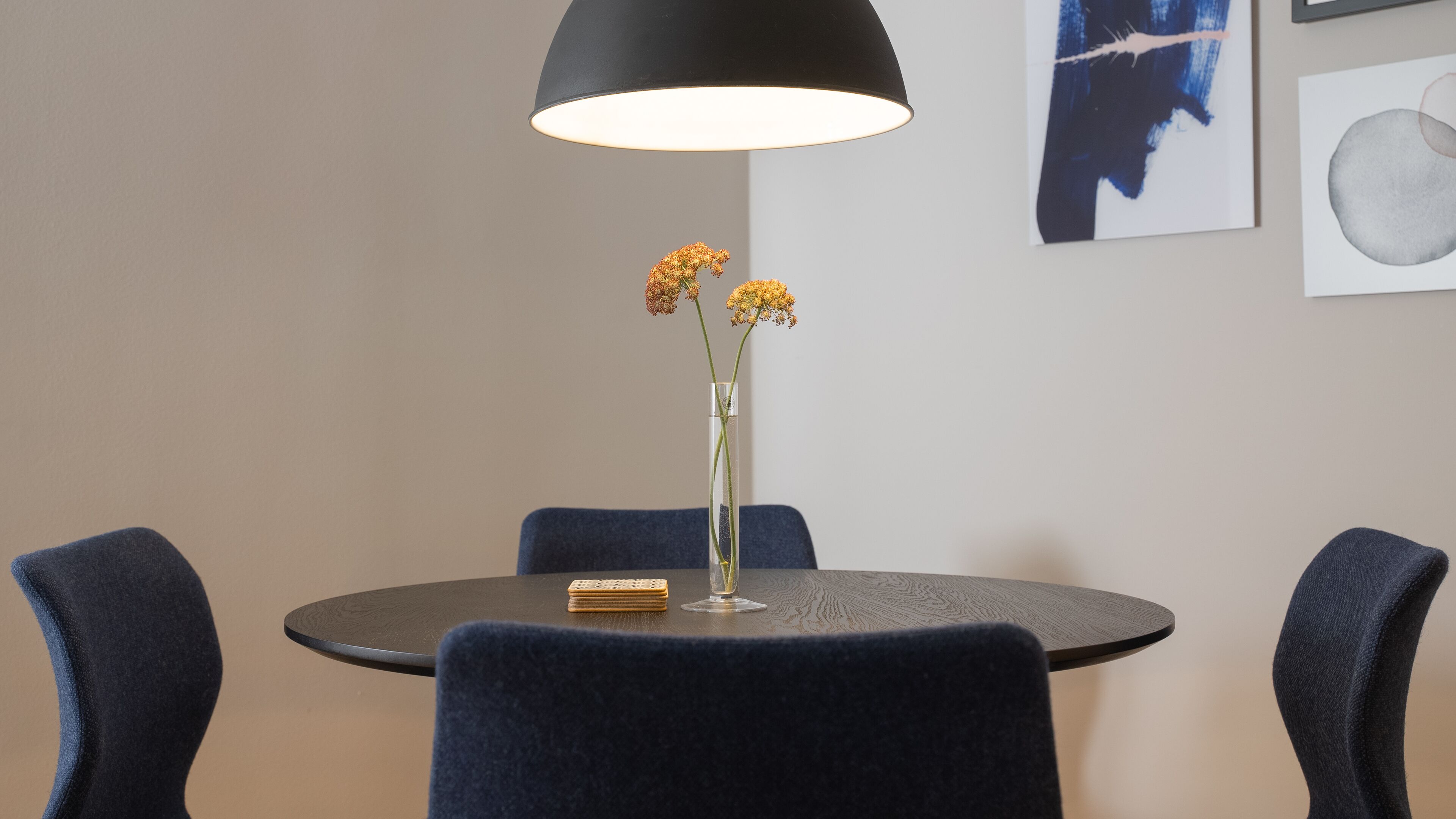 Une table ronde est agrémentée d’un vase haut et étroit contenant des fleurs jaunes, et est entourée de quatre chaises bleu foncé. Une lampe suspendue noire est fixée au-dessus de la table.