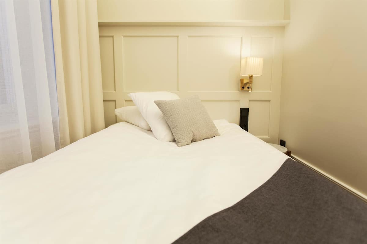 A neatly made bed with white linens and a grey throw blanket, a bedside lamp, and a window with sheer curtains.