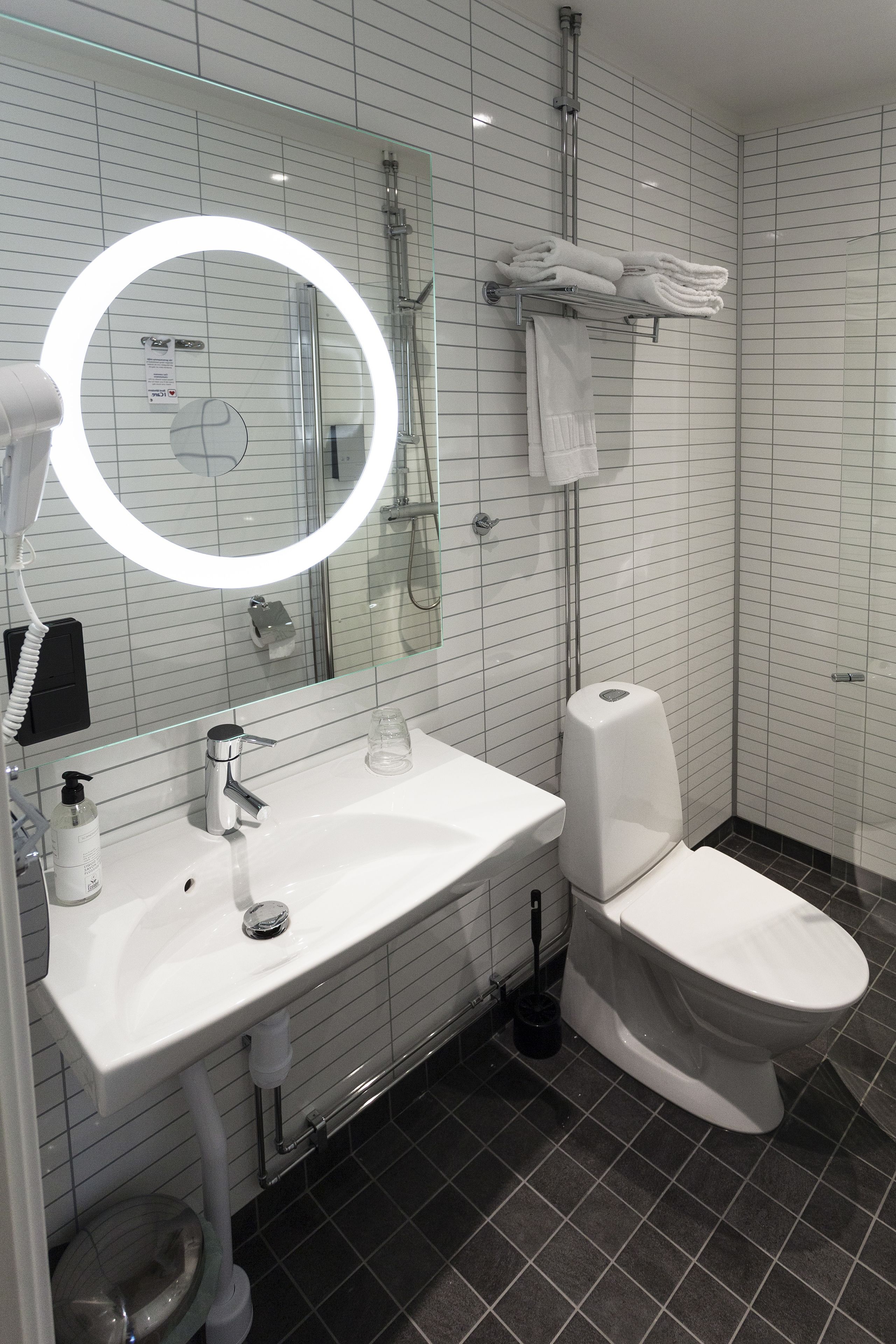 Un baño moderno con un lavabo blanco, un inodoro, una cabina de ducha de vidrio y un espejo redondo con luz.