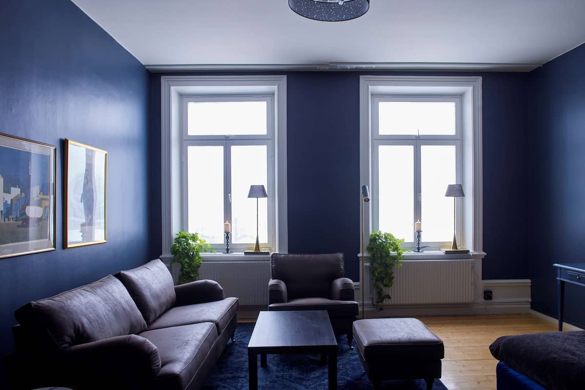 A living room with a dark blue wall, a brown leather sofa, a wooden coffee table, and two windows with lamps.