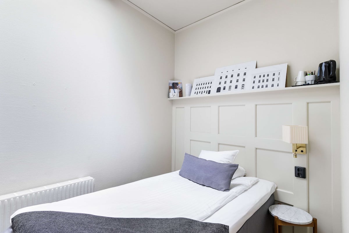 A neatly arranged bedroom with a bed, a small bedside table, a wall-mounted shelf with books and a lamp, and a radiator.